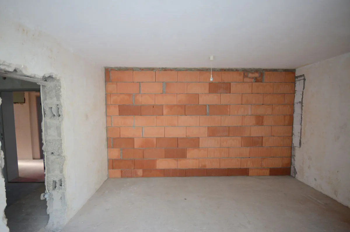Frontaler Blick auf eine unverputzte Ziegelwand im Rohbau mit Türöffnung links und einzelner Glühbirne an der Decke – vor der Modernisierung durch Broßler Küche Aktiv