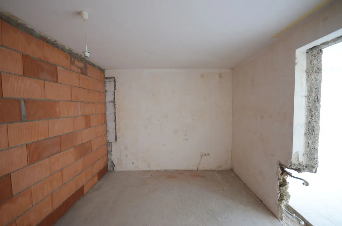 Rohbauraum mit massiver Ziegelwand auf der linken Seite, Fensterausschnitt rechts und gespachtelten Wänden – vor der Modernisierung durch Broßler Küche Aktiv