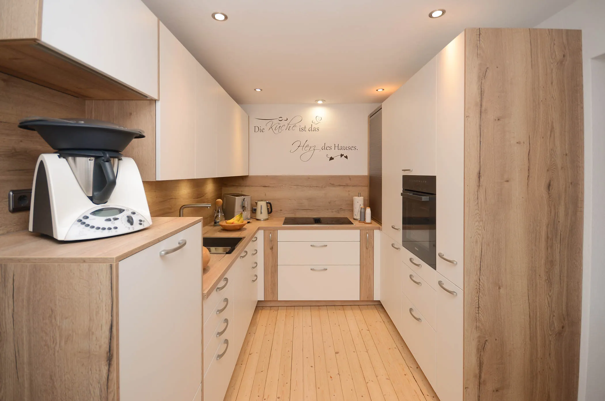 Moderne U-Küche mit weißen Fronten, Holz-Arbeitsplatte, Holz-Nischenrückwand mit Wandtattoo ‚Die Küche ist das Herz des Hauses‘, Backofen-Hochschrank in Holzoptik und Thermomix – Referenzprojekt von Broßler Küche Aktiv