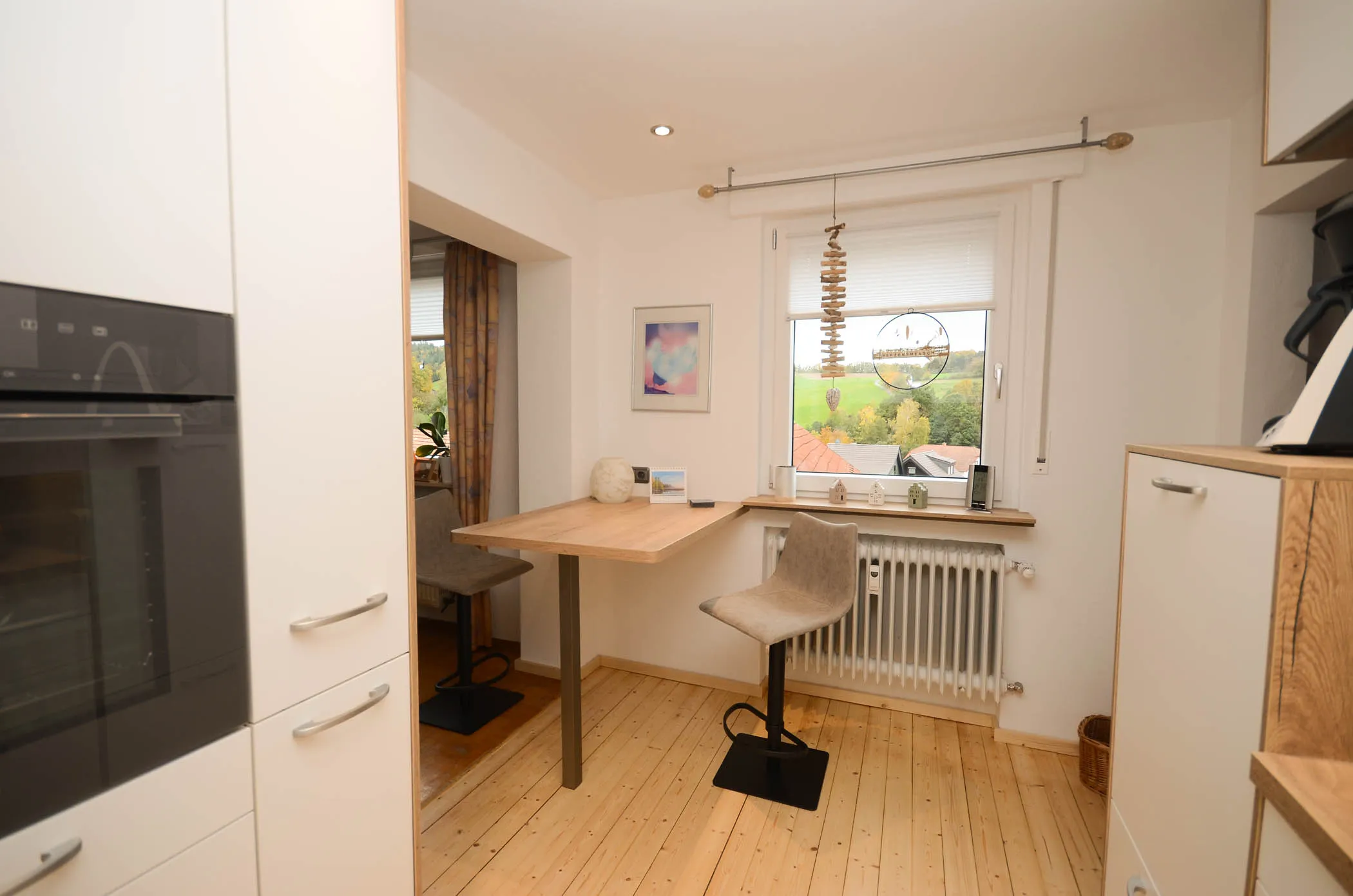 Helle Essecke in der neuen Küche mit Holztisch und zwei Polsterhockern vor dem Fenster, Backofen-Hochschrank links und Pendelleuchte über dem Tisch – Referenzprojekt von Broßler Küche Aktiv