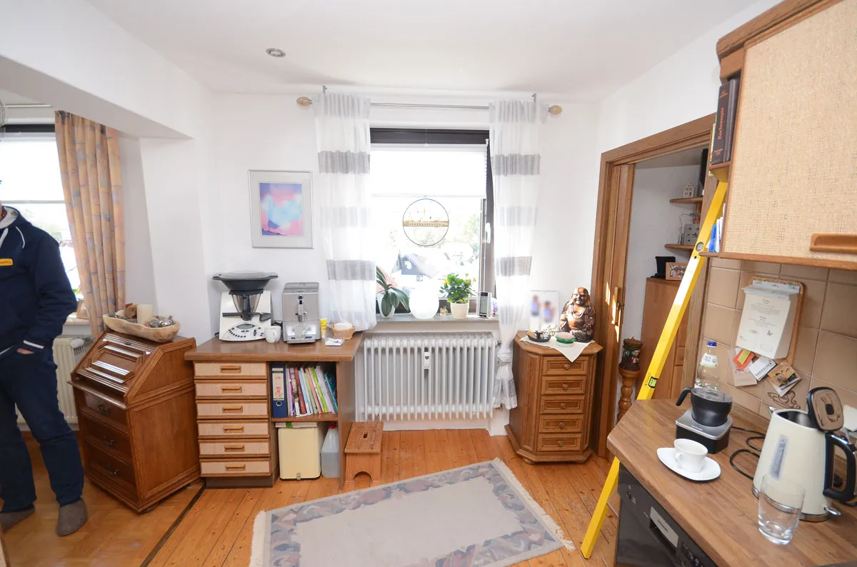 Bewohnter Wohnraum mit alten Holzmöbeln, Dielenboden, Heizkörper vor Fenster mit Gardinen und Bildern an der Wand – vor der Modernisierung durch Broßler Küche Aktiv