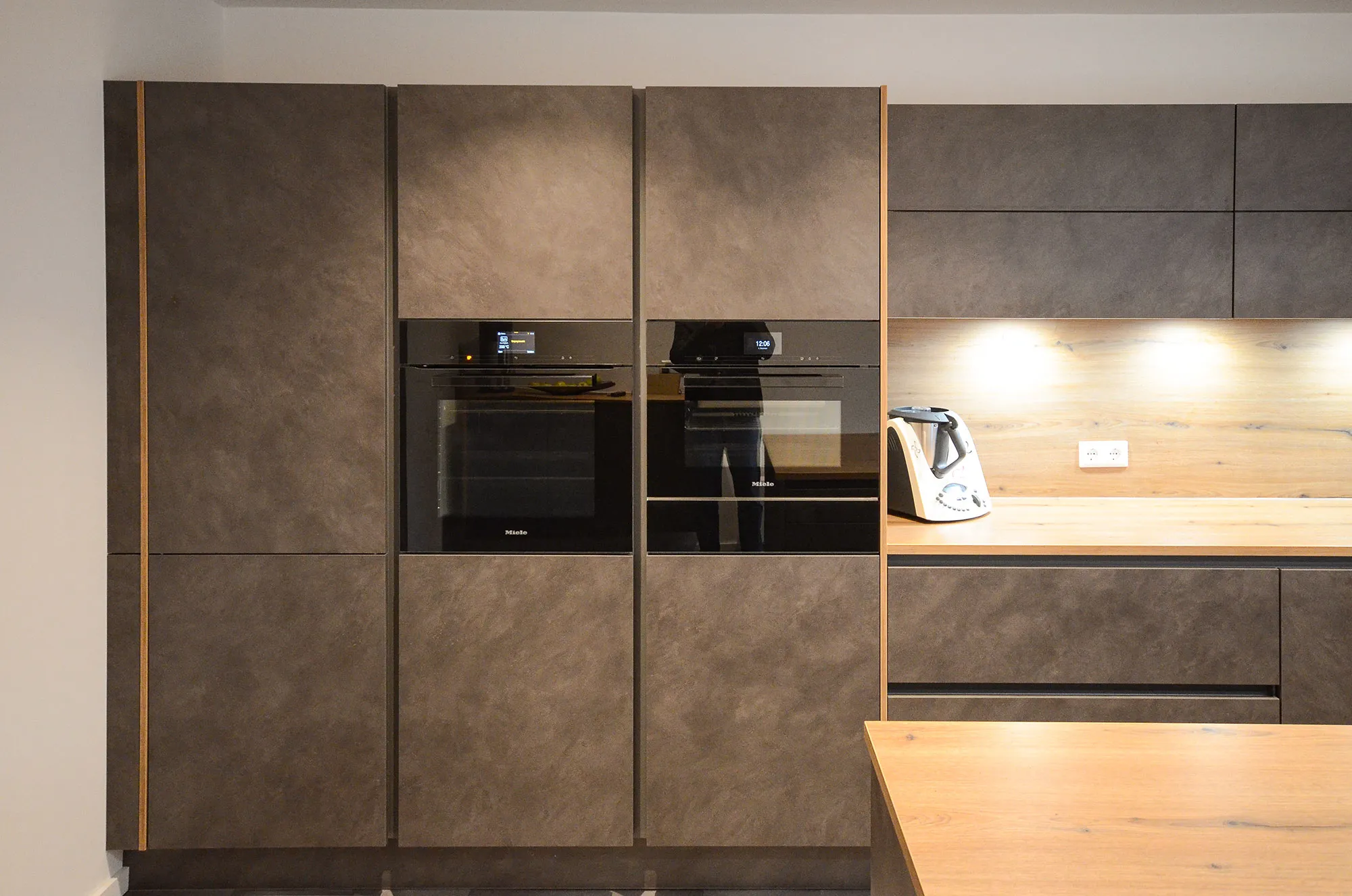Detailansicht der Schrankwand in dunkler Steinoptik mit übereinander integriertem Einbaubackofen und Dampfgarer, beleuchteter Holz-Nischenrückwand mit Thermomix und Halbinsel mit Eichen-Arbeitsplatte rechts – Referenzprojekt von Broßler Küche Aktiv