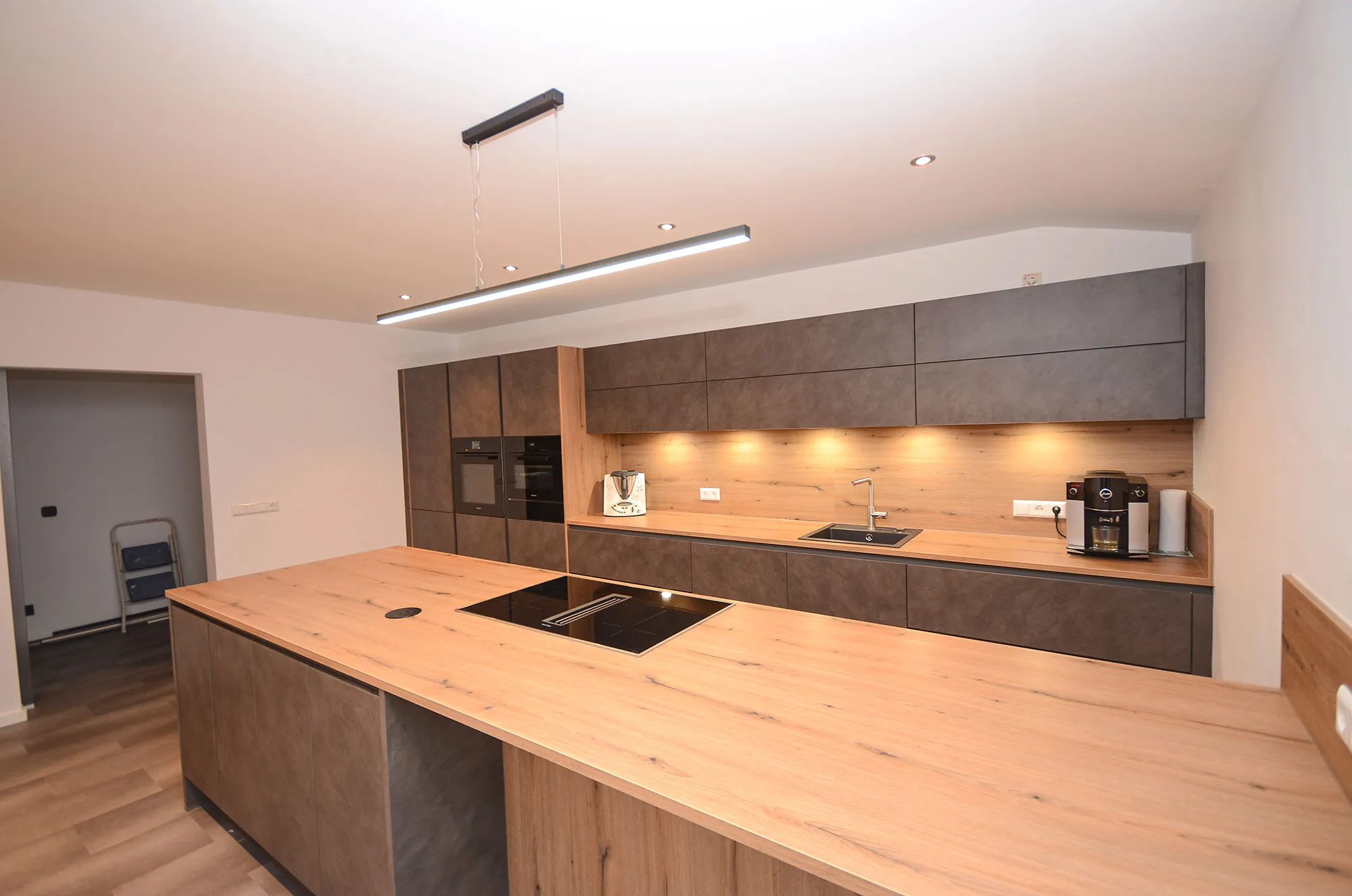 Frontale Längsansicht der modernen Küche mit Kochinsel mit Eichen-Arbeitsplatte und Kochfeld im Vordergrund, dahinter Schrankwand in dunkler Steinoptik mit beleuchteter Holz-Nischenrückwand, Spüle und Kaffeevollautomat, lineare Pendelleuchte über der Insel – Referenzprojekt von Broßler Küche Aktiv
