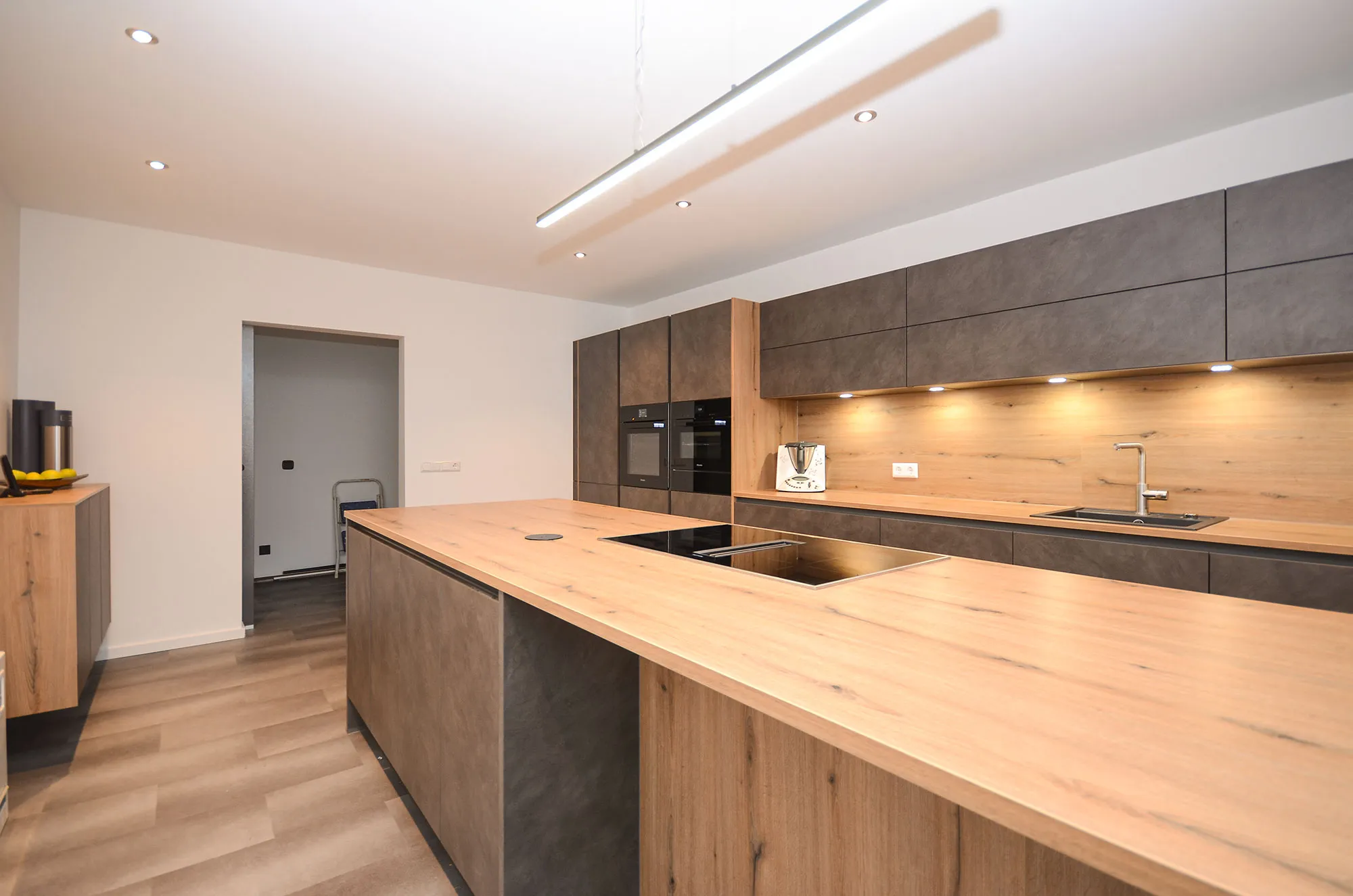Längsansicht einer modernen Küche mit großer Kochinsel mit Eichen-Arbeitsplatte und integriertem Kochfeld, dahinter Schrankwand in dunkler Steinoptik mit beleuchteter Holz-Nischenrückwand, Backofen-Türmen und Spüle, lineare Pendelleuchte über der Insel – Referenzprojekt von Broßler Küche Aktiv
