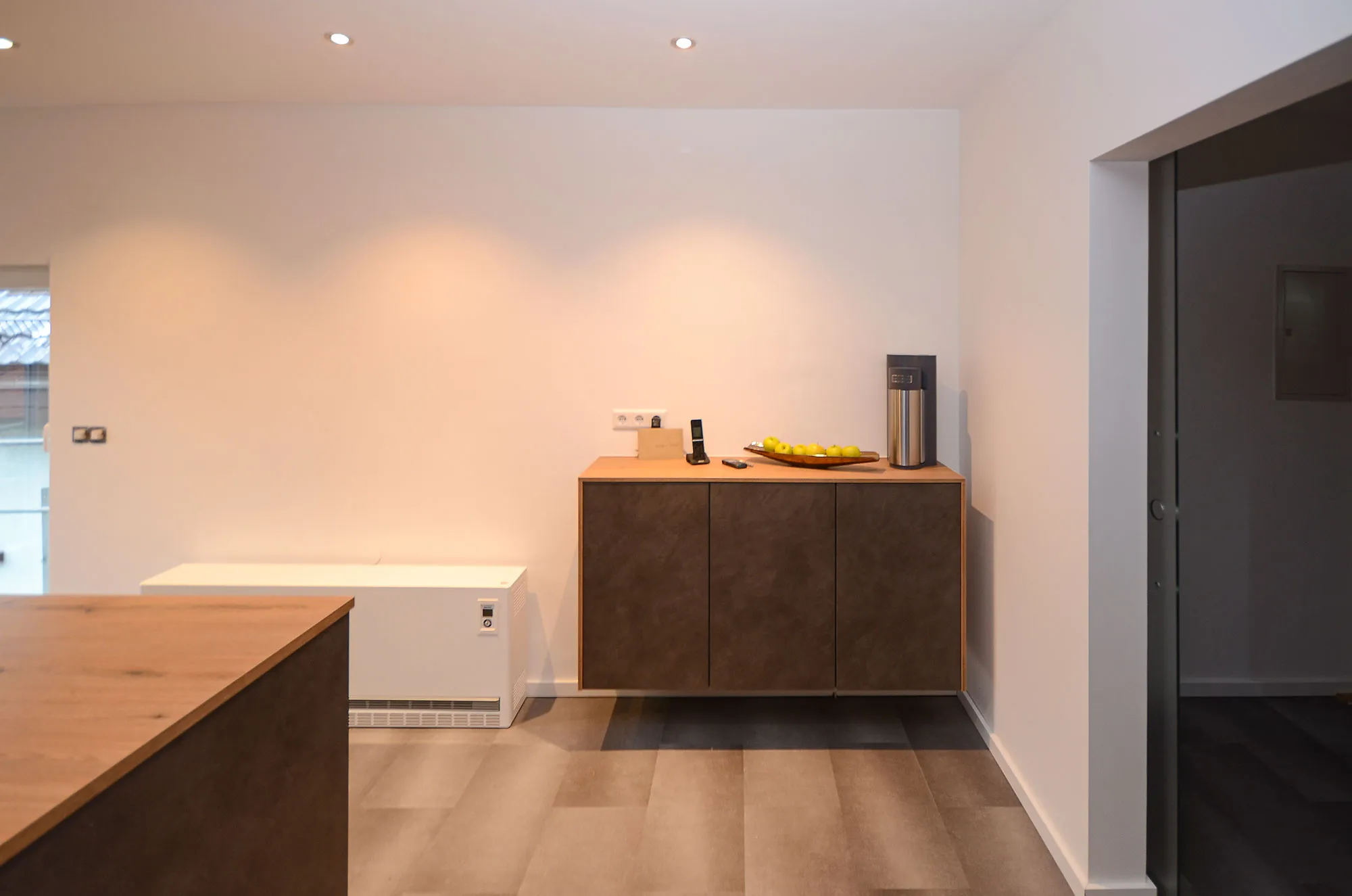 Sideboard in dunkler Steinoptik mit Obstschale und Telefon, dahinter eine weiße Kühltruhe und Holzboden, Ausblick auf die Kochinsel links – Referenzprojekt von Broßler Küche Aktiv
