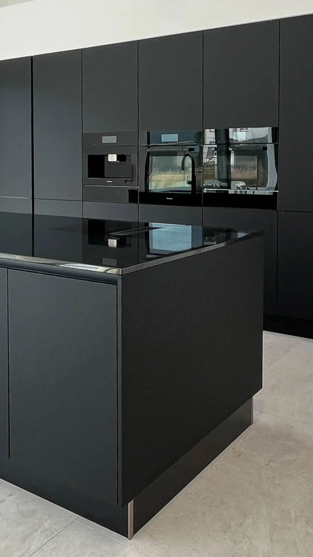 Schwarze grifflose Küche mit Kochinsel und schwarzer Steinarbeitsplatte, im Hintergrund Hochschrankzeile mit integriertem Backofen, Mikrowelle und Kaffeevollautomat in Schwarz, heller Steinfliesenboden – Küchenplanung von Dietmar Kraus für Broßler Küche Aktiv