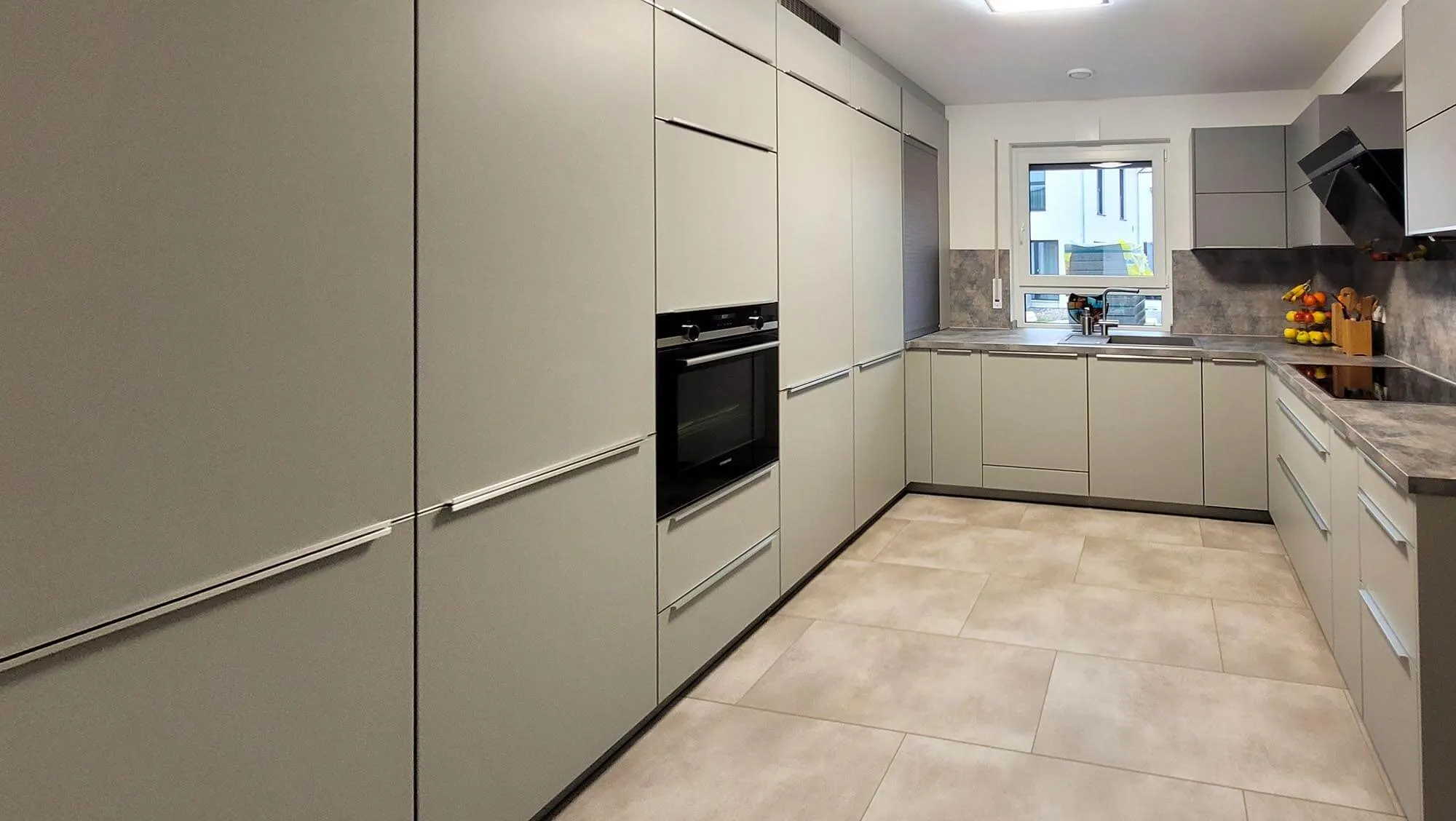 Gesamtansicht der beigen L-Küche mit Hochschrankwand links inkl. eingebautem Backofen und Tambour-Schrank, rechts Arbeitszeile mit Spüle am Fenster und Induktionskochfeld