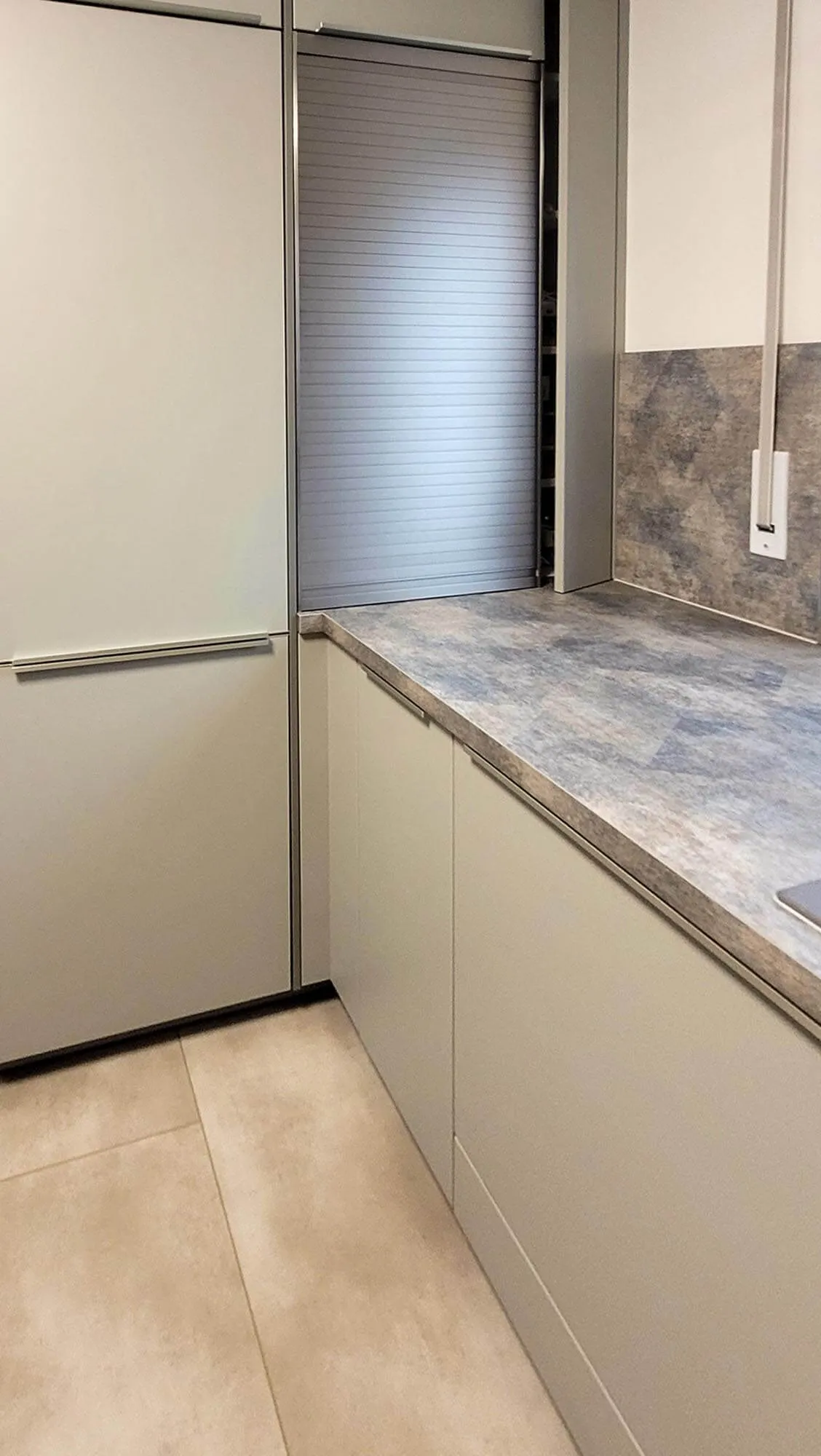 Eckbereich der Küche mit Tambour-Aufsatzschrank, beigen grifflosen Unterschränken und Steinoptik-Arbeitsplatte über Eck, Anschluss für Wand-Dunstabzug