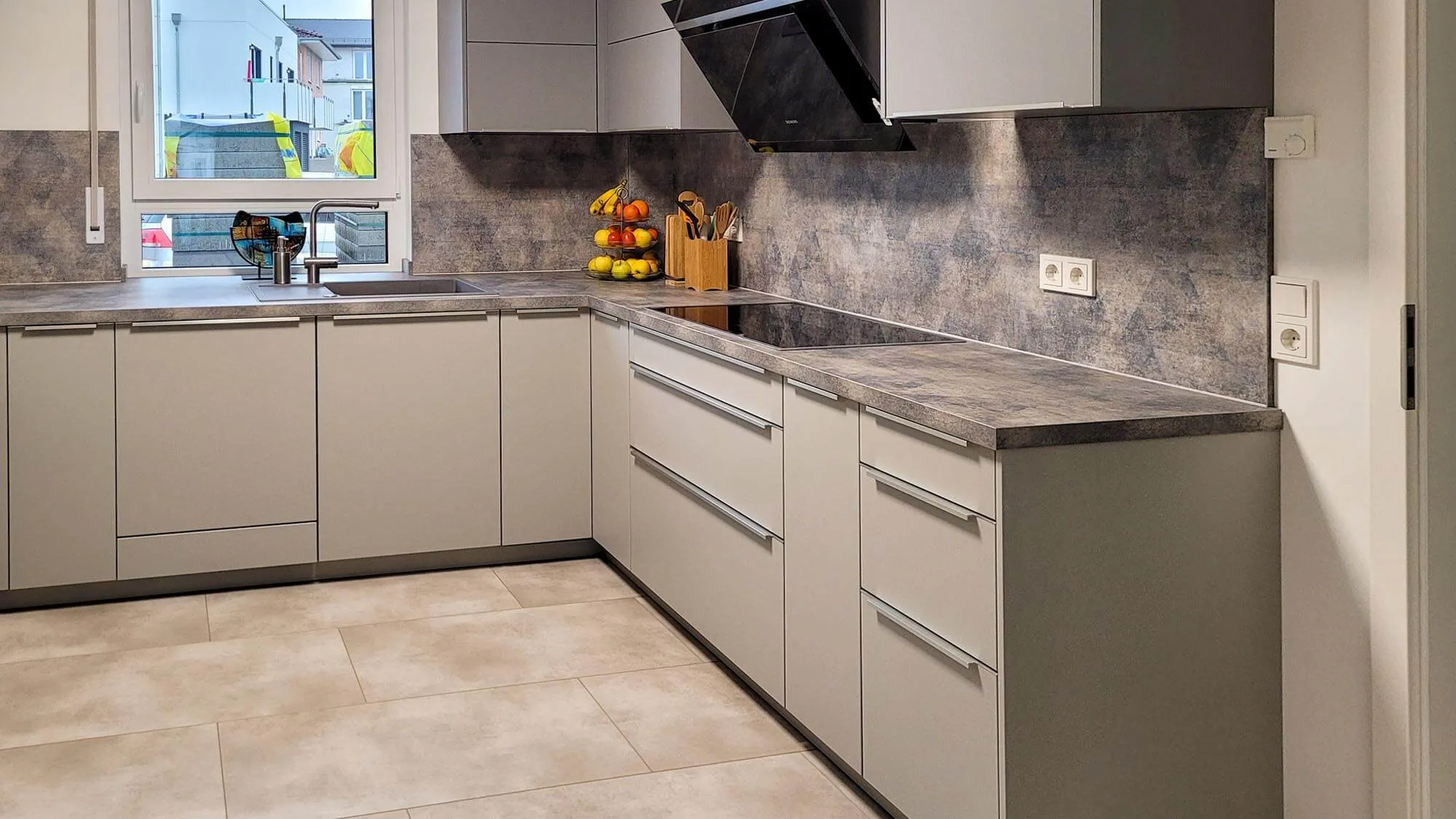 Helle L-Küche mit beigen grifflosen Fronten, Steinoptik-Arbeitsplatte und Rückwand, Induktionskochfeld mit Wand-Dunstabzug und Spüle am Fenster