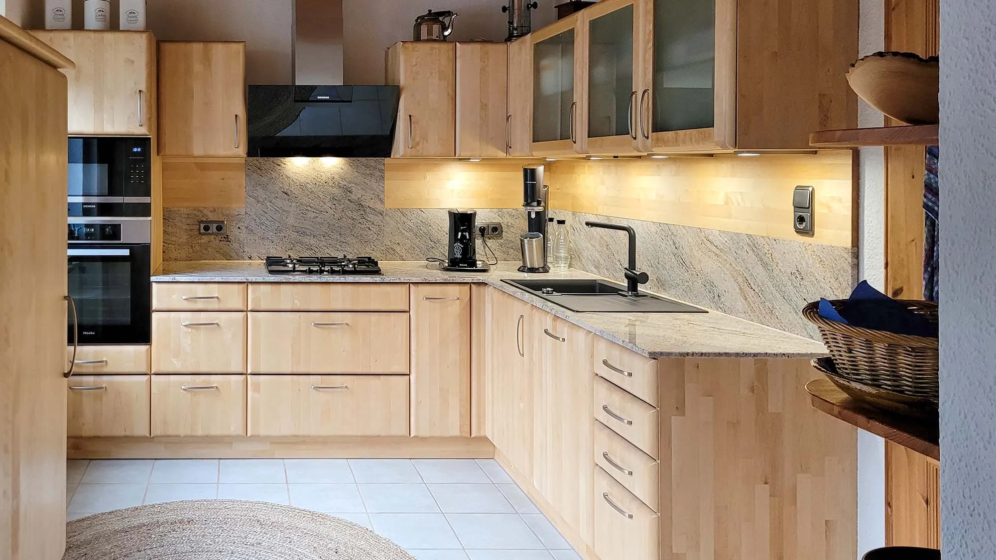 Helle L-Küche aus heller Eiche mit Granit-Arbeitsplatte und passender Rückwand, Glas-Hängeschränken, Backofen-Hochschrank links, Gaskochfeld und Spüle