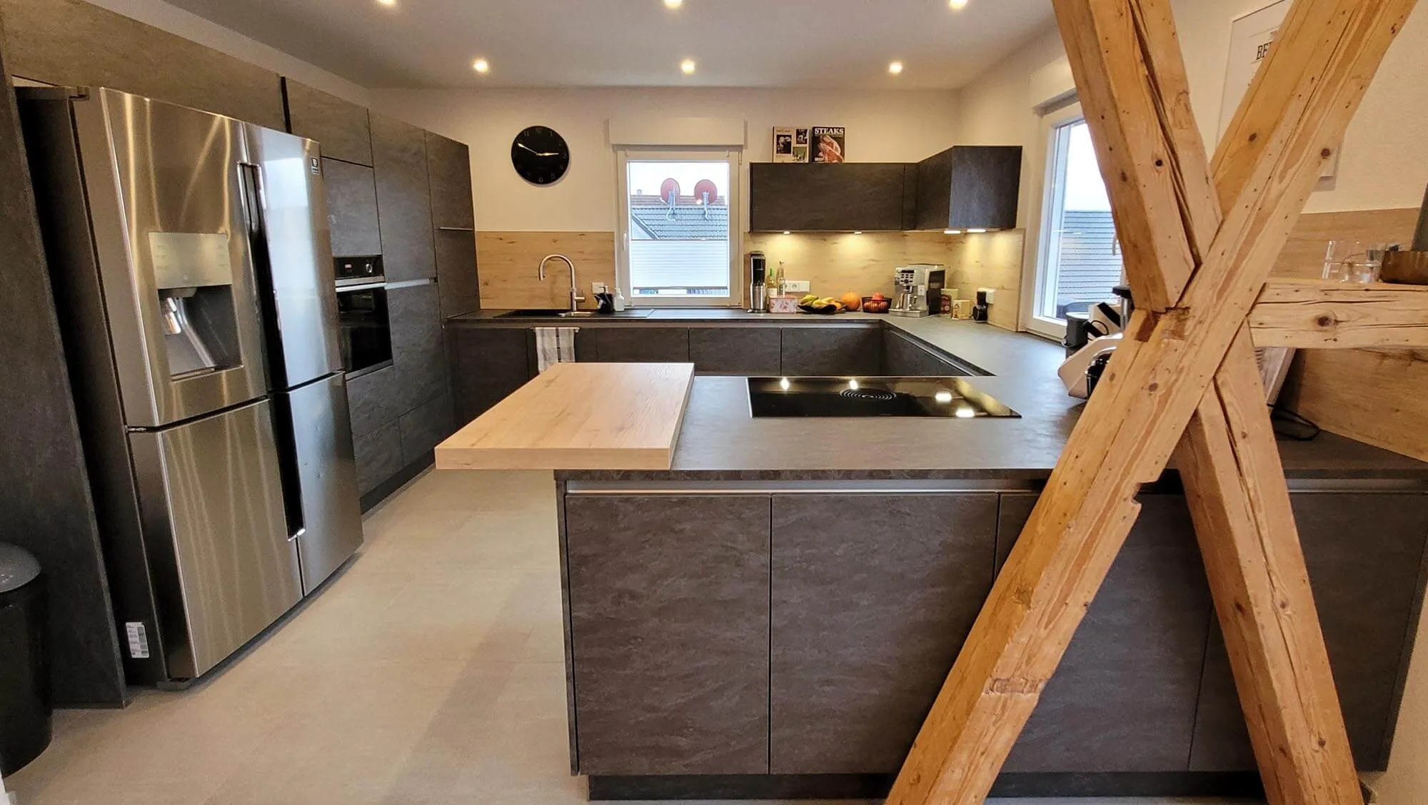 Gesamtansicht der dunkelbraunen U-Küche im Dachgeschoss mit Halbinsel und Holz-Bartheke, Side-by-Side-Edelstahlkühlschrank, Backofen-Hochschrank und im Vordergrund markante Holzbalken-X-Konstruktion
