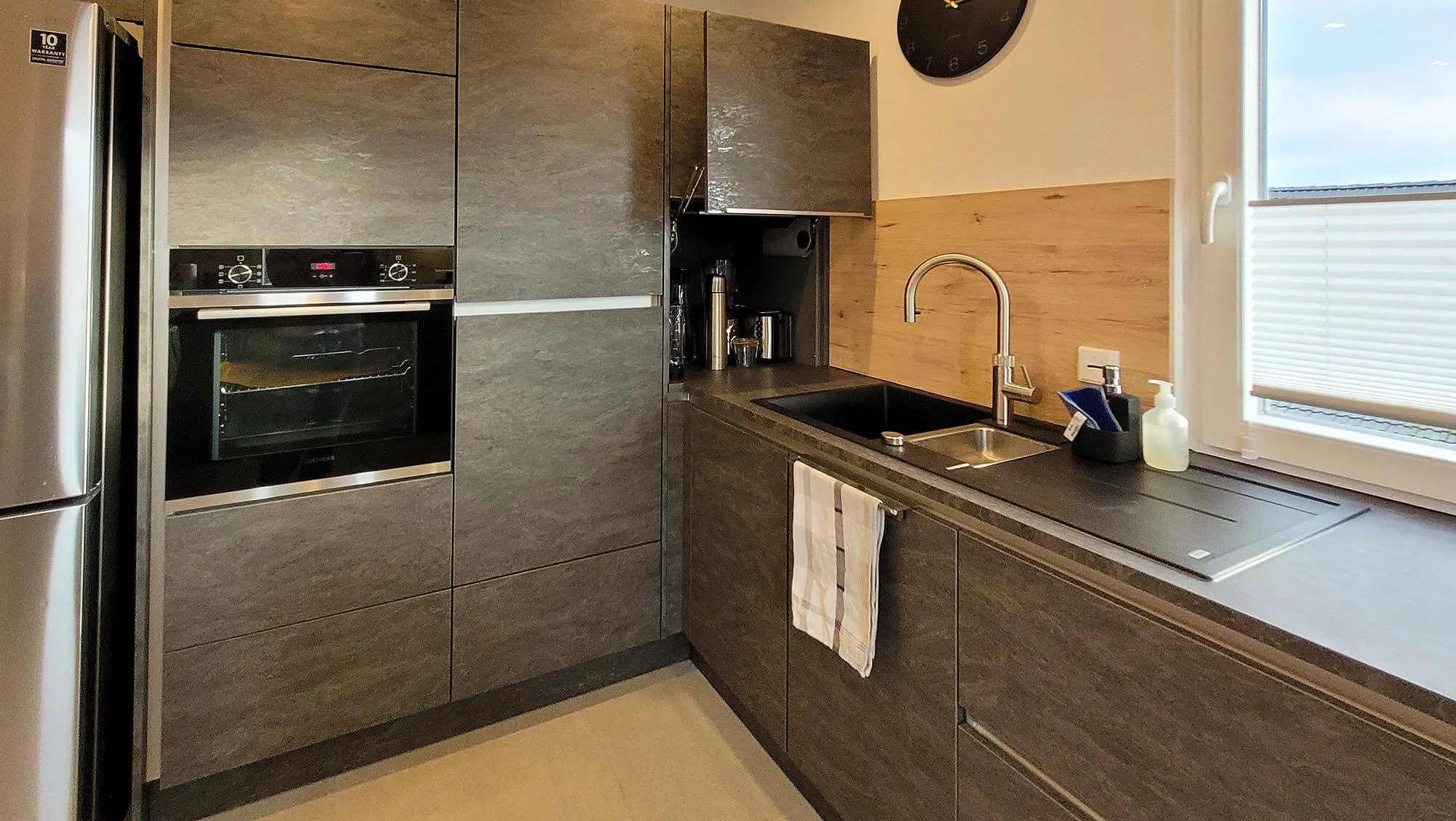 Detail Spülbereich mit Bogenarmatur, integrierter Abtropffläche in der Arbeitsplatte, Holz-Spritzschutz über Eck und Backofen-Hochschrank in dunkler Holzoptik