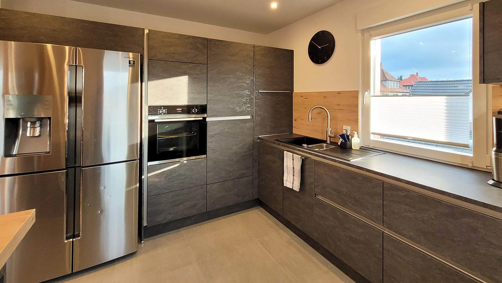 Arbeitszeile mit Side-by-Side-Edelstahlkühlschrank, dunklem Backofen-Hochschrank und Spüle am Fenster mit Holz-Spritzschutz, schwarzer Wanduhr und grifflosen Fronten in Dunkelbraun