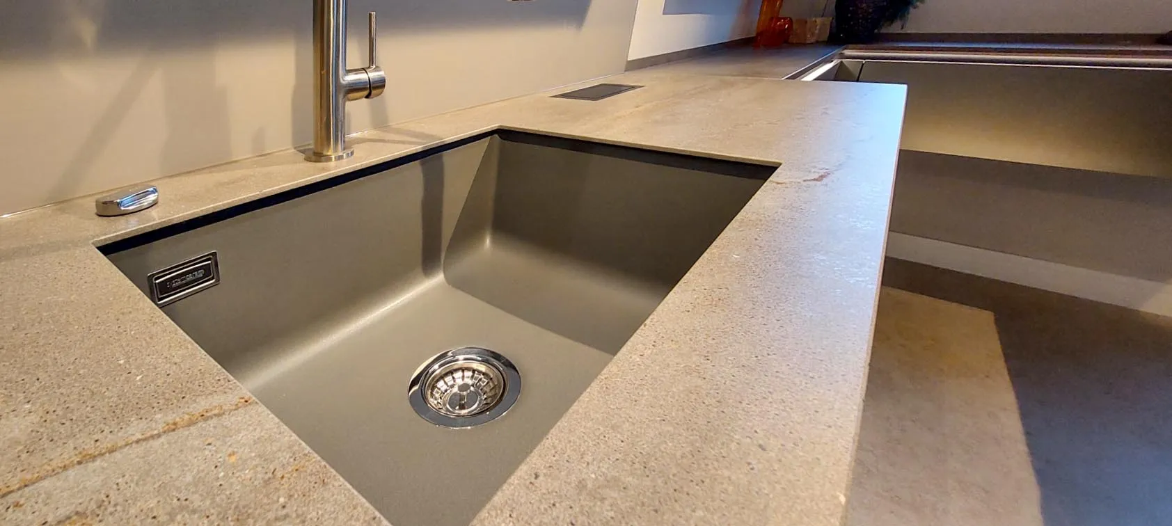 Detail Spülbereich mit großer Unterbau-Edelstahlspüle, Schiebebetatigung des Ablaufs und Sockel-Armatur in einer dunkelgrauen Steinarbeitsplatte