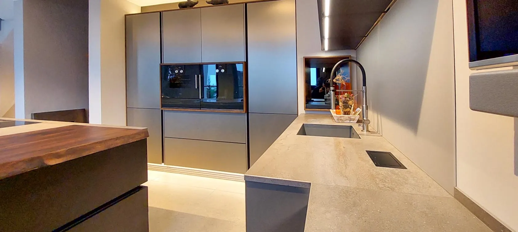 Innenansicht der Küche mit Hochschrankzeile, Backofen und Side-by-Side-Kühlschrank, gegenüber Arbeitszeile mit Spüle und Bogenarmatur, links Holz-Bartheke der Kochinsel, mittig Holznische