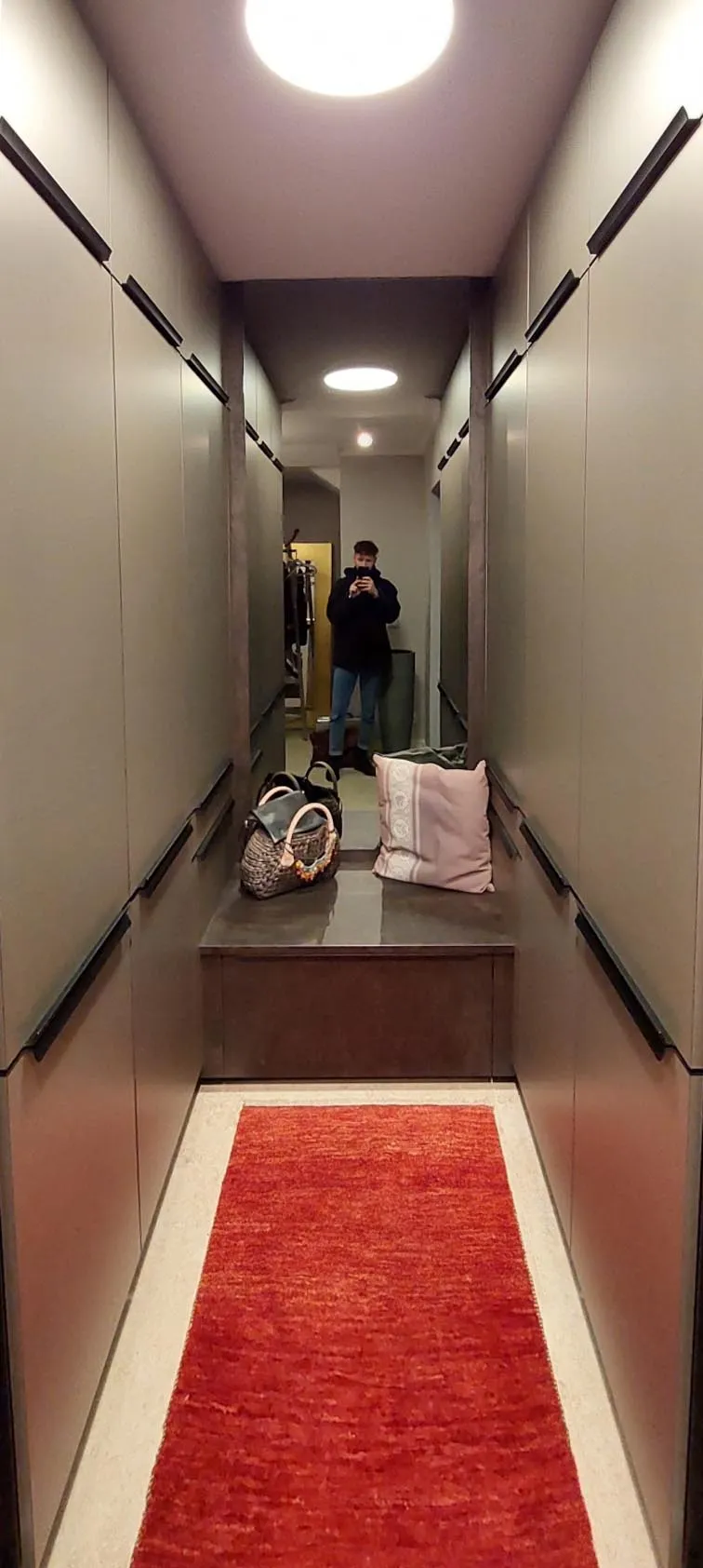 Vorher-Aufnahme eines schmalen begehbaren Ankleidezimmers mit beidseitigen Hängeleisten, Holz-Sitzbank am Ende und Spiegel, am Boden roter Läufer und Taschen