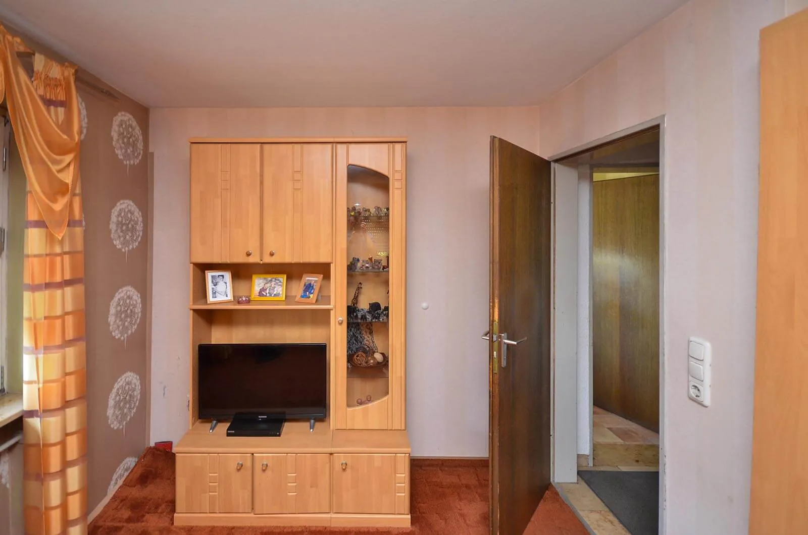 Vorher-Aufnahme des angrenzenden Wohnraums im bewohnten Zustand mit massivem Kiefernholz-Wohnzimmerschrank, Röhren-Fernseher, gemustertem Teppich und Vorhängen aus 1990er-Jahren, Blick durch eine offene Tür in den Flur – Bestandssituation vor Umbau und Küchenplanung von Dietmar Kraus bei Broßler Küche Aktiv