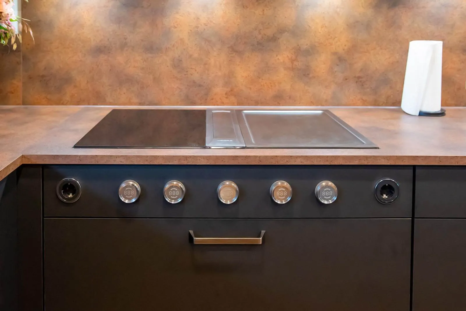 Nahaufnahme des Gas-Kochfelds mit fünf Edelstahl-Drehknöpfen und integriertem Teppan-Yaki-Element auf kupferfarbener Arbeitsplatte, anthrazitfarbene Kochinsel-Front mit Bronzegriff, Rückwand in passender Patina-Optik – Detail einer Küchenplanung von Dietmar Kraus für Broßler Küche Aktiv