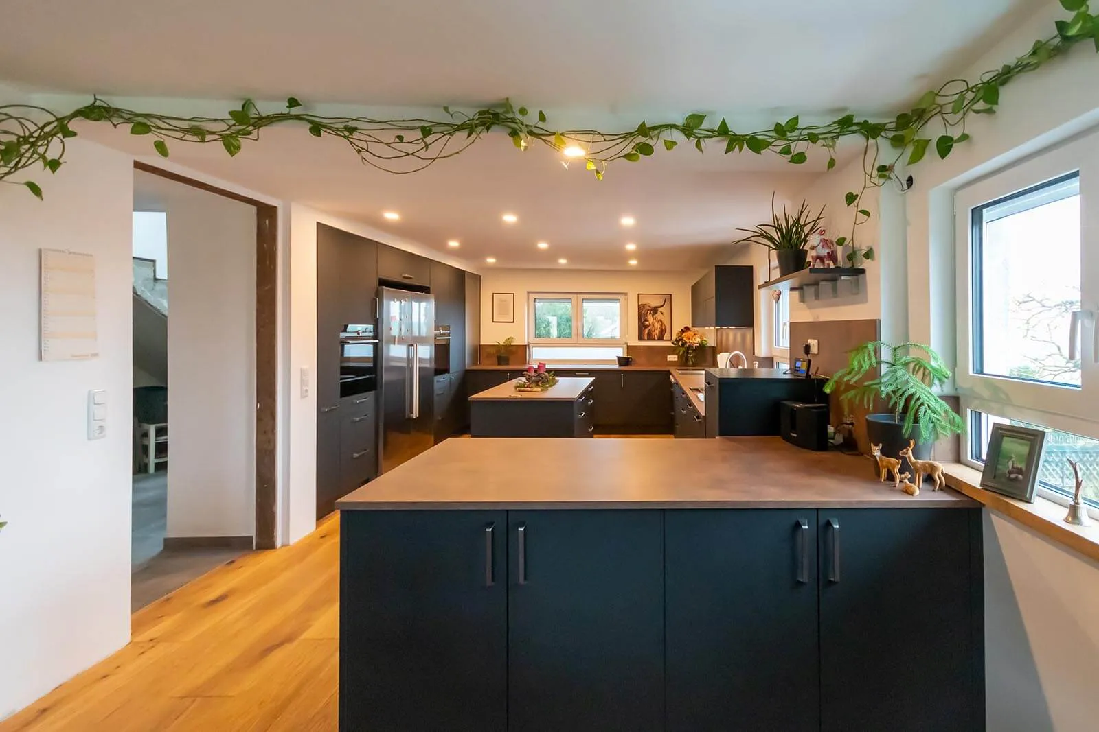 Moderne grifflose Küche in mattem Anthrazit mit massiver Arbeitsplatte in warmer Eichen-Holzoptik, großzügige Kochinsel mit Sitztheke, integrierter Edelstahl-Kühlschrank und Backofenturm, Eichendielen-Boden und hängende Grünpflanzen über der Insel – Küchenplanung von Dietmar Kraus für Broßler Küche Aktiv