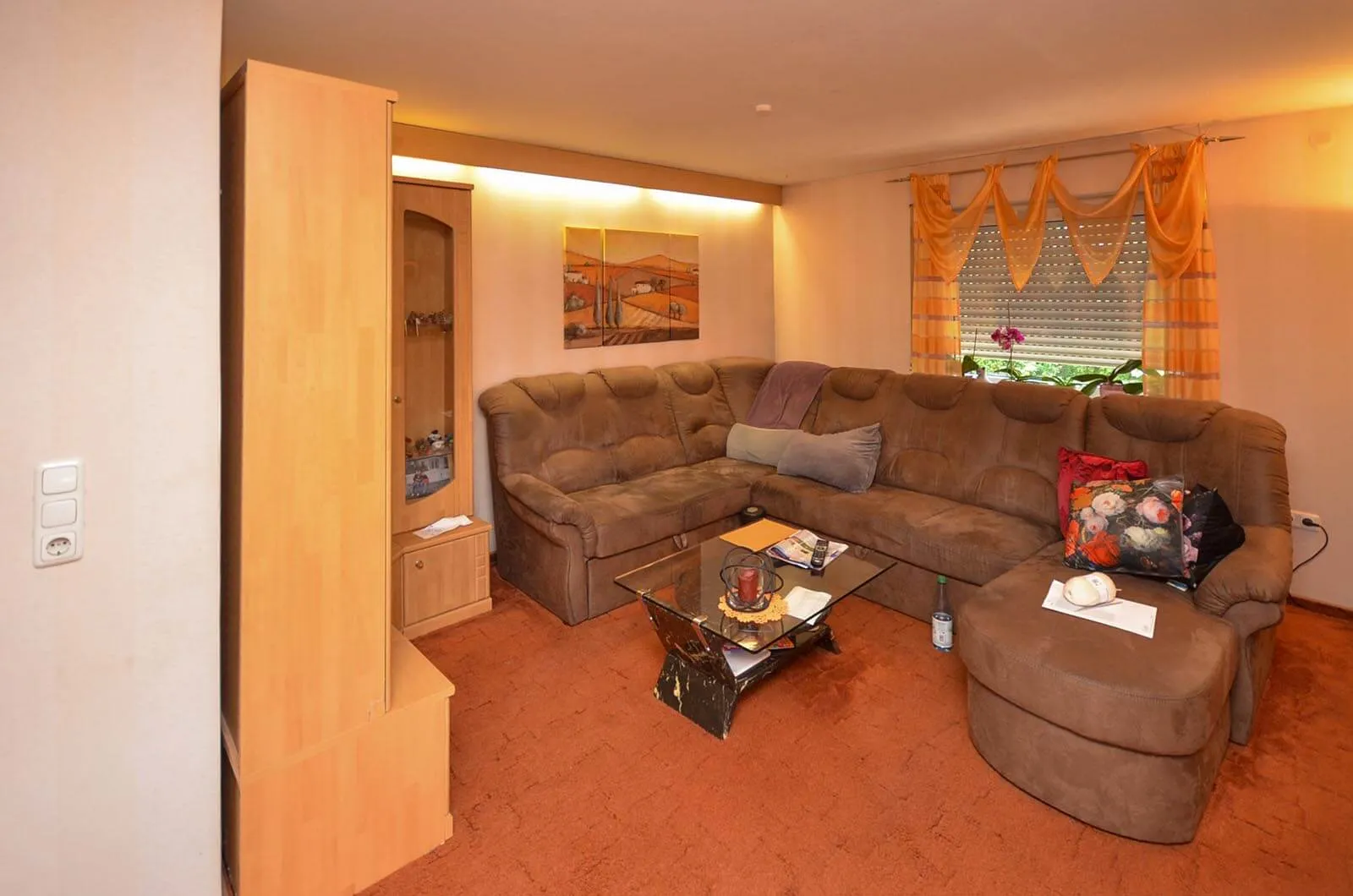 Vorher-Aufnahme des Wohnraums aus weiterer Perspektive mit großem braunem Cord-Ecksofa, Glas-Couchtisch, oranger Teppichboden und Vorhängen, links Vitrine mit Glasaufsatz und Wandbild – Bestandssituation vor Umbau und Küchenplanung von Dietmar Kraus bei Broßler Küche Aktiv