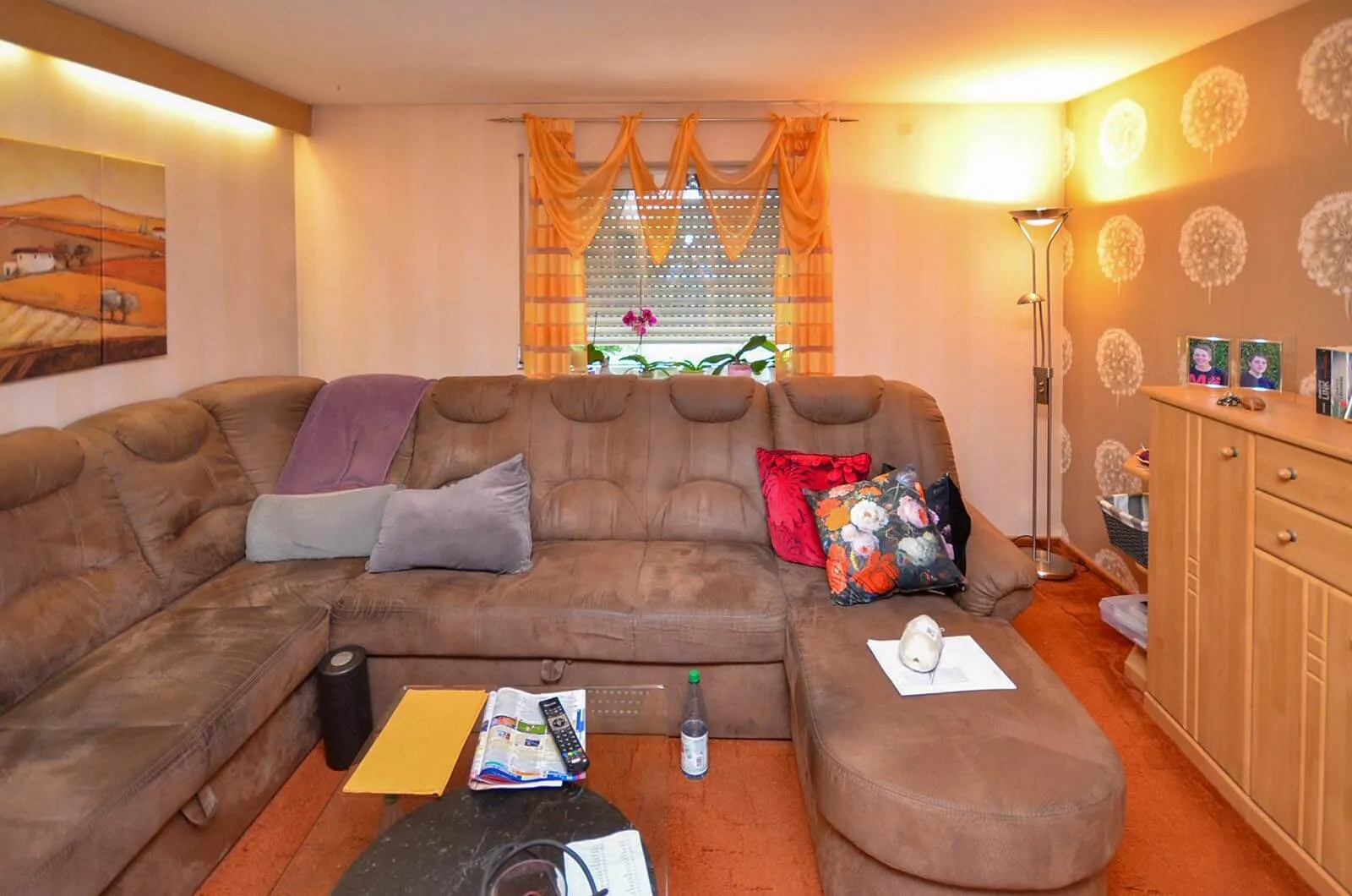 Vorher-Aufnahme desselben Wohnraums aus anderer Perspektive mit braunem Cord-Ecksofa, lila Kissen, oranger Teppichboden, orangefarbene Vorhänge, Kiefernholz-Sideboard rechts, Stehlampe und gemusterte Tapete – Bestandssituation vor Umbau und Küchenplanung von Dietmar Kraus bei Broßler Küche Aktiv