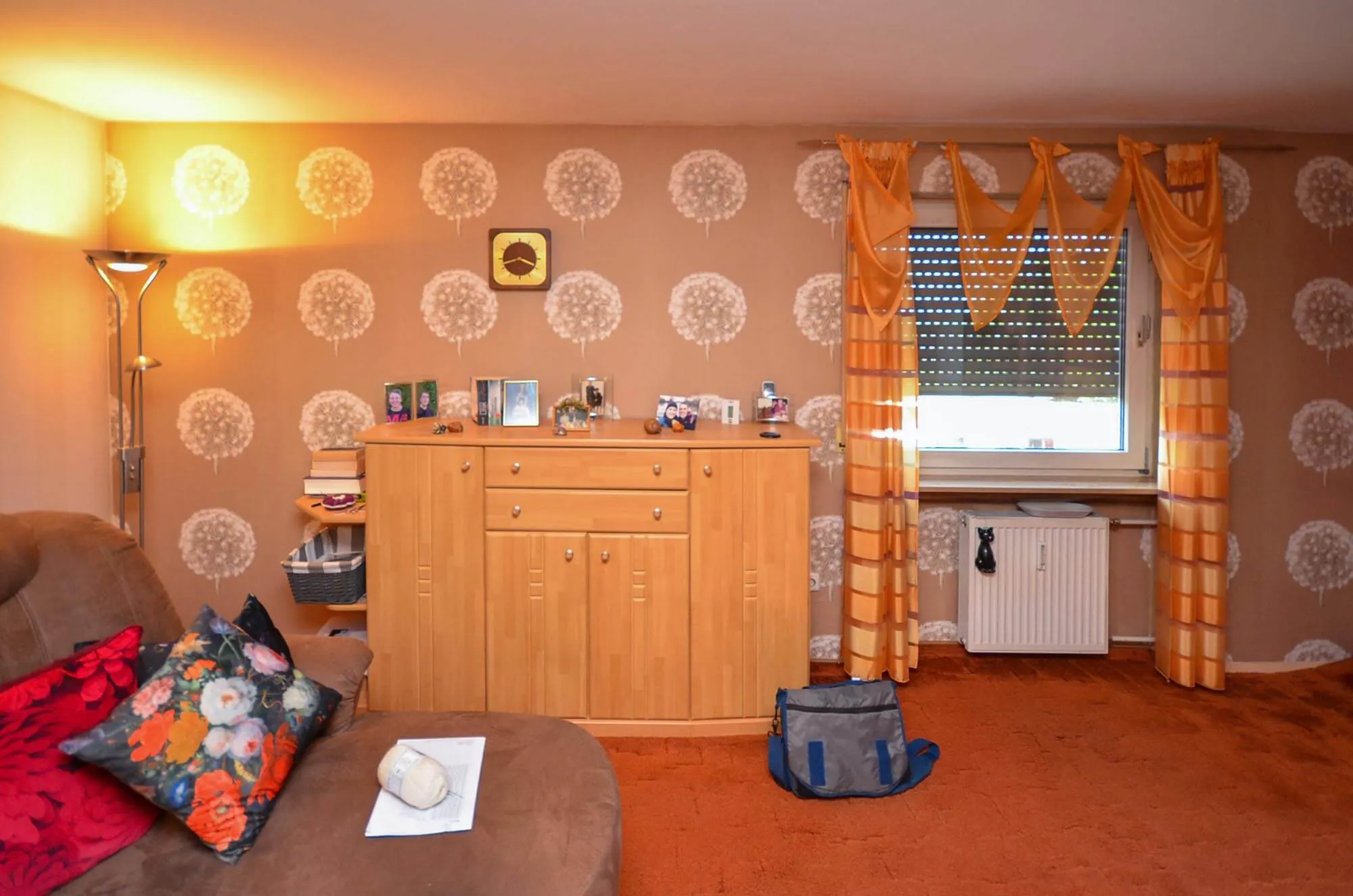 Vorher-Aufnahme eines Wohnraums im bewohnten Zustand mit gemusterter Tapete und passenden orangefarbenen Vorhängen, Kiefernholz-Sideboard, oranger Teppichboden und braunes Sofa mit Blumenkissen, Stehlampe links – Bestandssituation vor Umbau und Küchenplanung von Dietmar Kraus bei Broßler Küche Aktiv