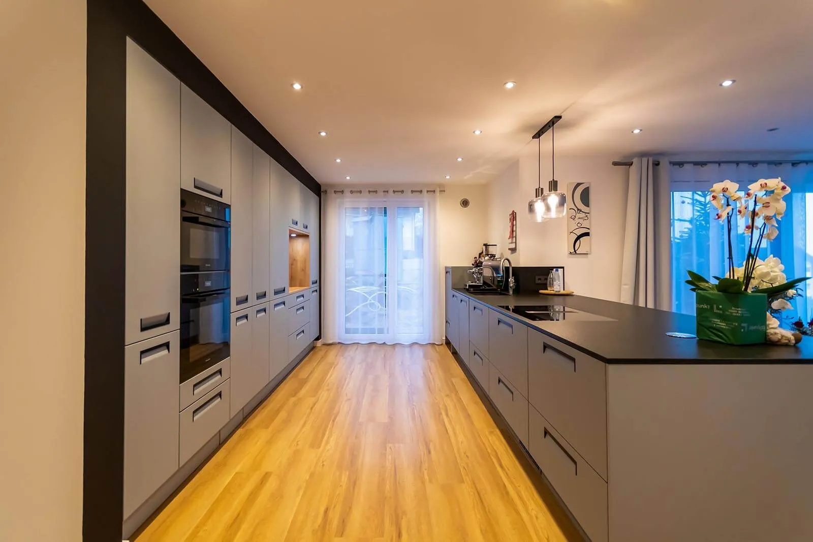 Langgestreckte Küche in beige-grauer Mattoptik mit schwarzer Arbeitsplatte, lange Kochinsel rechts mit Spüle und Orchideen-Deko, gegenüberliegende Hochschrankzeile mit integriertem Backofen, schwarze Pendelleuchten und Eichendielen-Boden, große Terrassentür im Hintergrund – Küchenplanung von Dietmar Kraus für Broßler Küche Aktiv