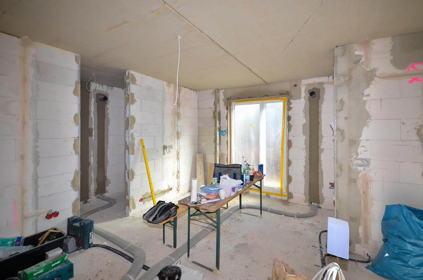 Vorher-Aufnahme eines frühen Neubau-Stadiums vor dem Kücheneinbau, unverputzte Porenbeton-Wände, gelb gerahmtes Fenster mit Folie, Tisch mit Werkzeug und Baumaterial im Raum, frische Rohinstallationen für Strom und Wasser – Ausgangssituation für eine Küchenplanung von Dietmar Kraus bei Broßler Küche Aktiv
