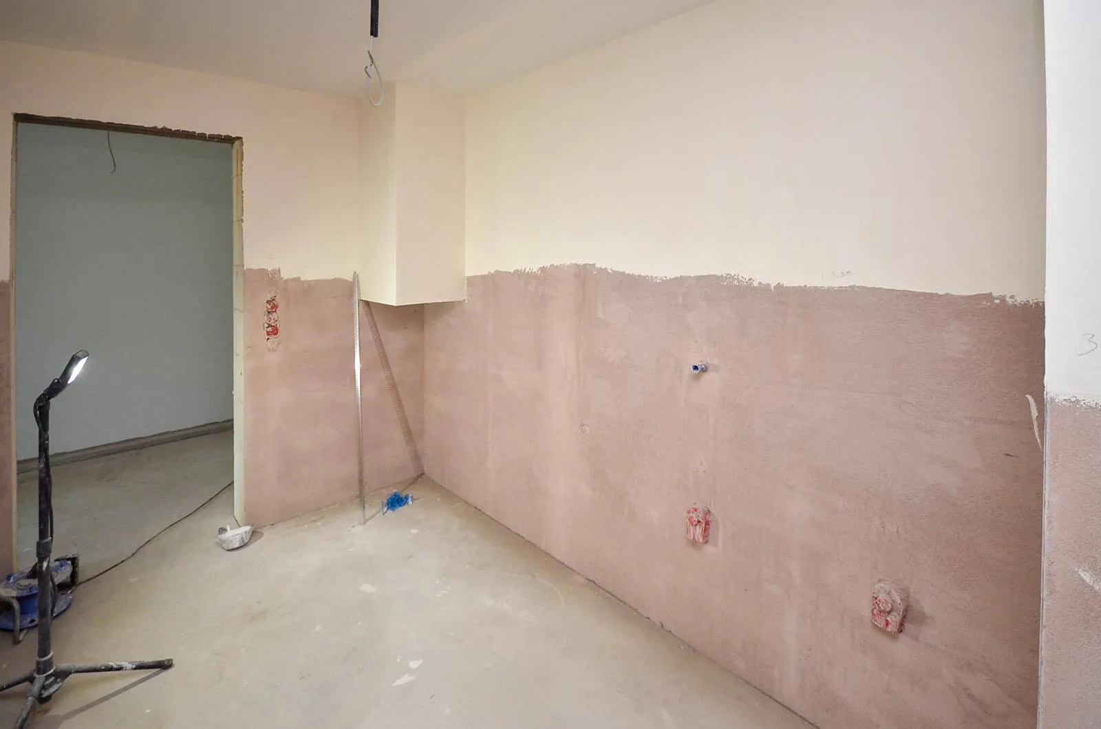 Vorher-Aufnahme einer leeren Raumecke im Rohbau vor dem Kücheneinbau, frisch verputzte Wände mit rosa Grundierung, sichtbare Vorinstallationen für Strom und Wasser, Estrich-Boden mit Baustellenlampe und Werkzeug – Ausgangssituation für eine Küchenplanung von Dietmar Kraus bei Broßler Küche Aktiv