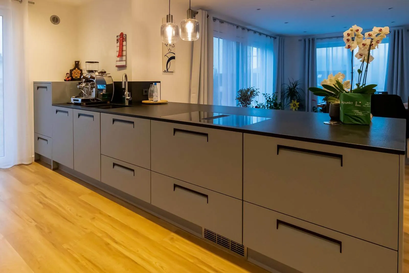 Lange grifflose Kochinsel in dezenter Beige-Mattoptik mit schwarzer Steinarbeitsplatte und integriertem Induktionskochfeld mit Muldenlüfter, schwarze Pendelleuchten und Espressomaschine im Hintergrund, Orchidee als Akzent, Eichendielen-Boden – Küchenplanung von Dietmar Kraus für Broßler Küche Aktiv