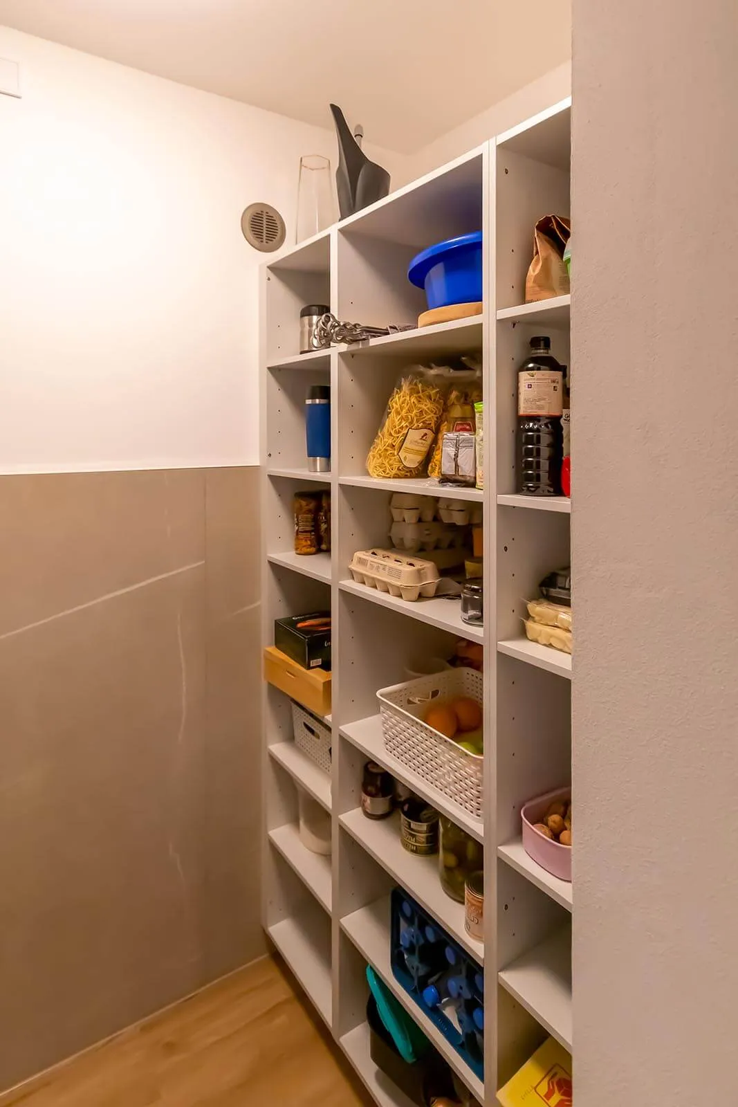 Vollständig bestücktes weißes Regal in der Speisekammer, sechs Ebenen mit Lebensmitteln, Getränken, Eierkartons, Trockenvorräten und Körben, beigefarbene Wandfliesen in Marmoroptik, Eichendielen-Boden – Planung von Dietmar Kraus für Broßler Küche Aktiv