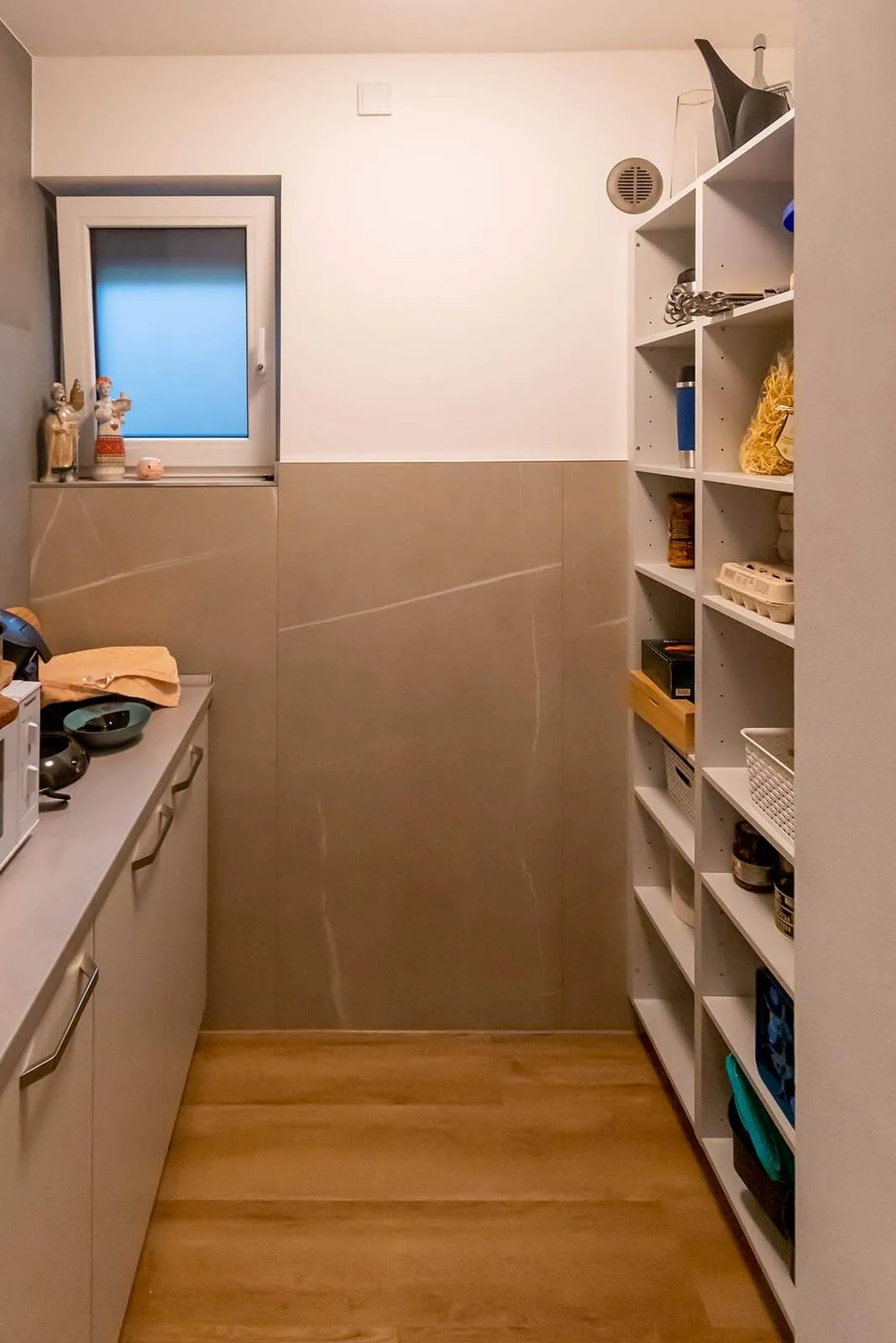 Speisekammer im Hochformat mit beigefarbenen Wandfliesen in Marmoroptik, offenes weißes Regal rechts mit Vorräten, kleines Fenster im Hintergrund, Eichendielen-Boden – Planung von Dietmar Kraus für Broßler Küche Aktiv