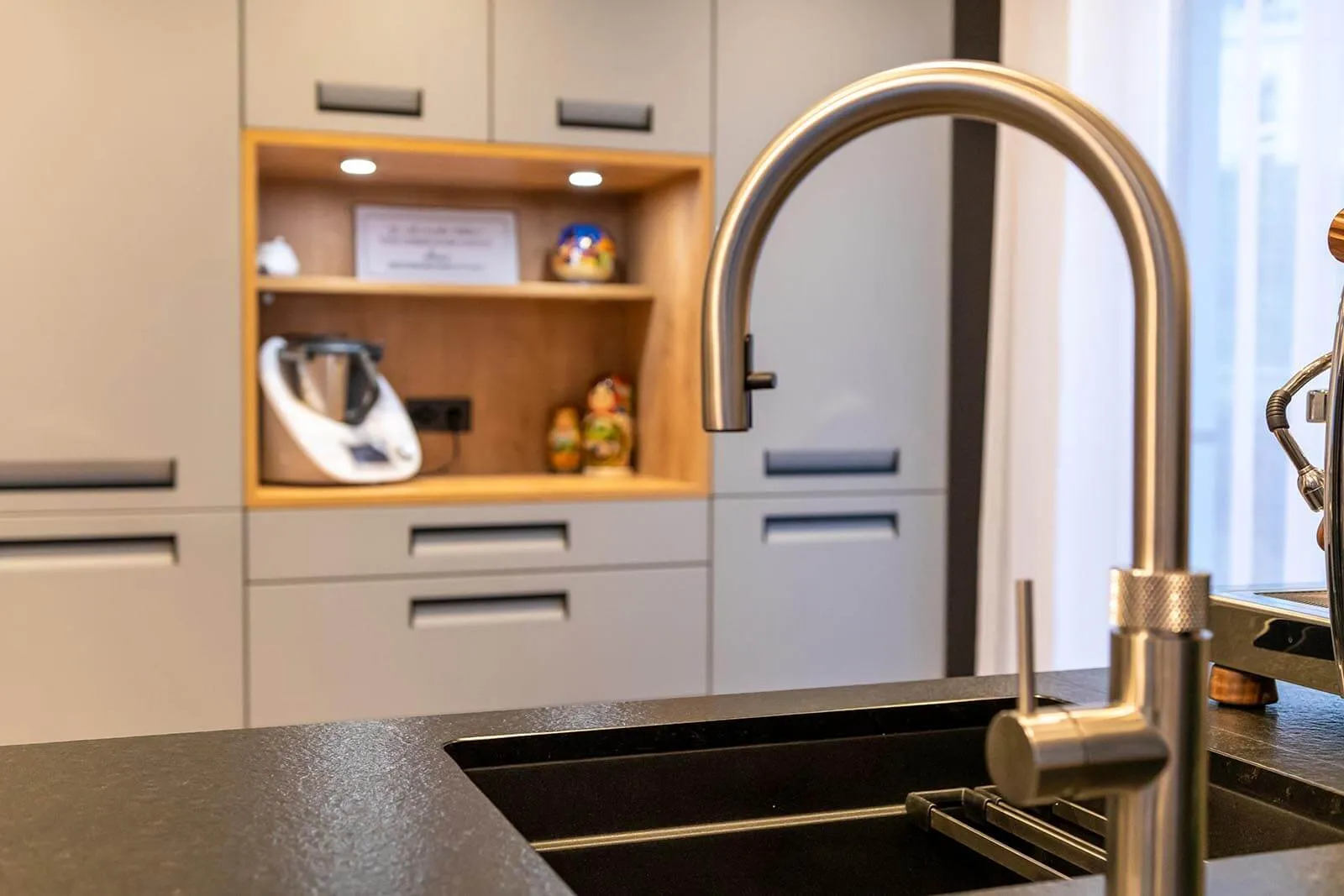 Nahaufnahme einer hohen Bronze-Armatur über einer Edelstahl-Unterbauspüle in schwarzer Steinarbeitsplatte, im Hintergrund unscharf eine offene Eichenholz-Nische mit Thermomix und Gewürzen – Detail einer Küchenplanung von Dietmar Kraus für Broßler Küche Aktiv