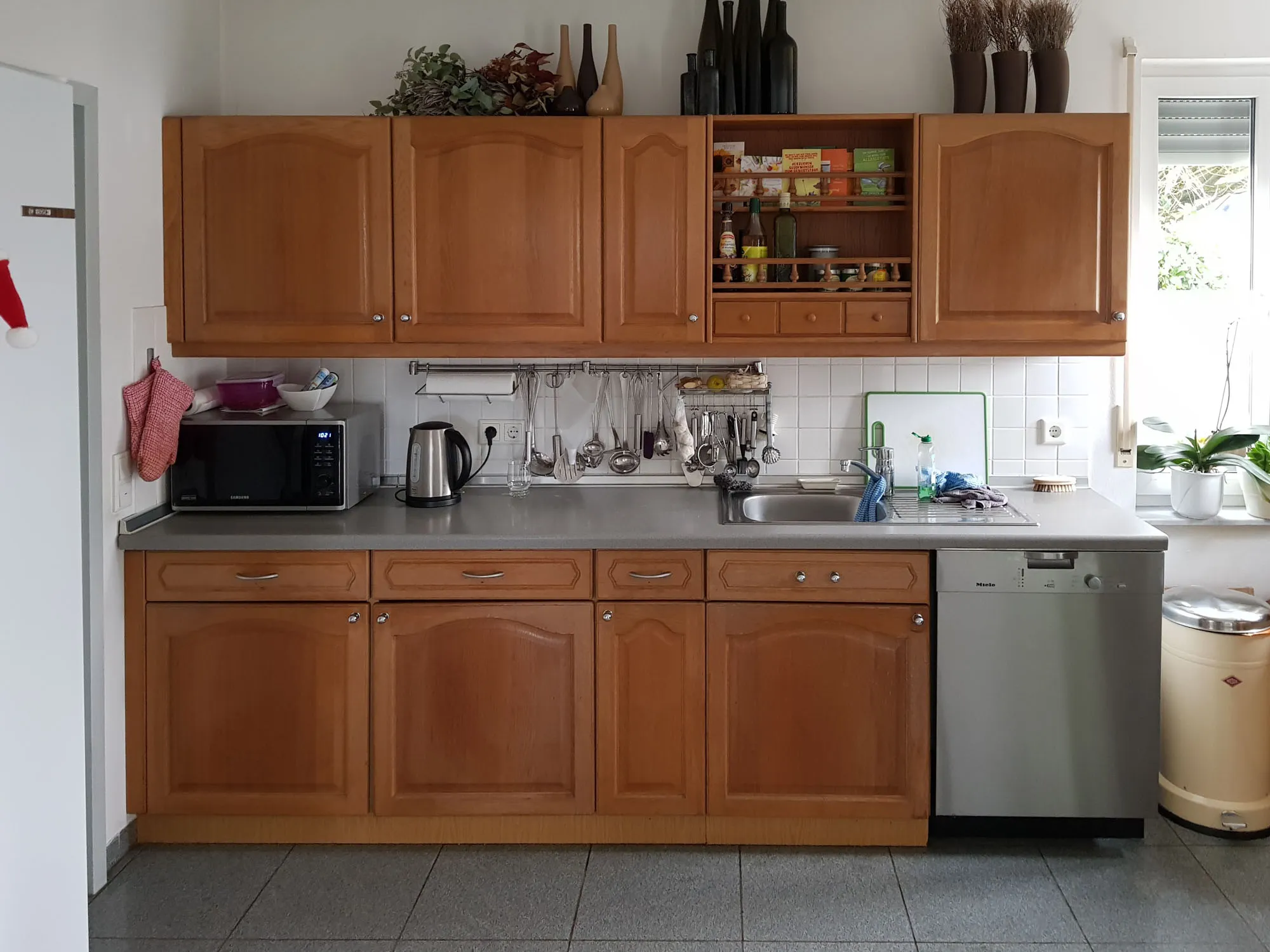 Vorher-Aufnahme der Spülen-Zeile der Bestands-Küche im Landhausstil mit Eichenholz-Kassettenfronten, Edelstahl-Spüle, integrierter Spülmaschine, Mikrowelle und Wasserkocher auf der Arbeitsplatte, weiße Wandfliesen, beigefarbener Treteimer rechts – Ausgangssituation vor der Küchenplanung von Dietmar Kraus bei Broßler Küche Aktiv