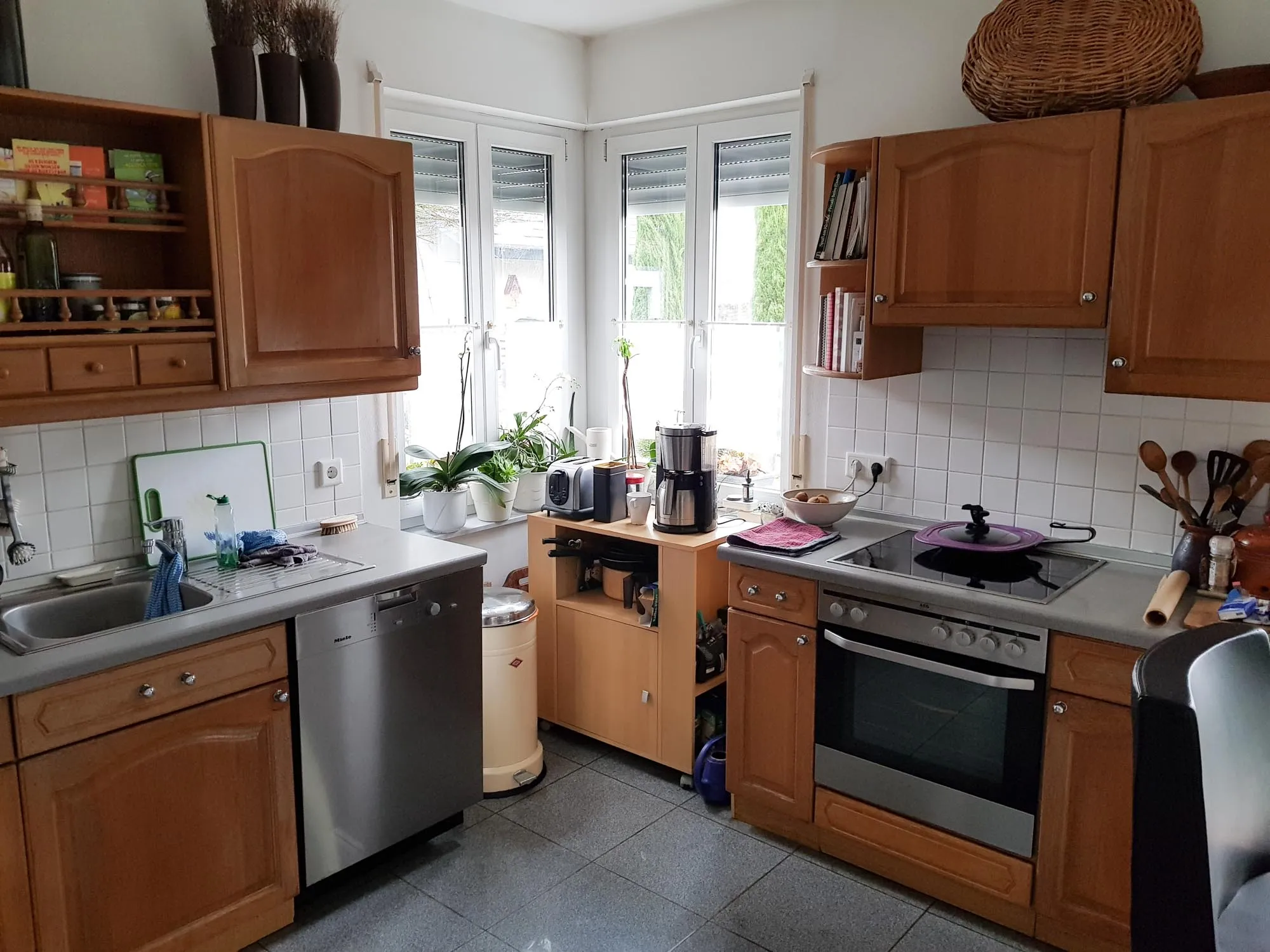 Vorher-Aufnahme einer Bestands-Küche im Landhausstil mit Eichenholz-Kassettenfronten, eingebauten Elektrogeräten (Spülmaschine, Backofen, Kochfeld), weißen Wandfliesen, dreiteiligem Fenster mit viel Tageslicht, Pflanzen und Küchenutensilien im bewohnten Zustand – Ausgangssituation vor der Küchenplanung von Dietmar Kraus bei Broßler Küche Aktiv