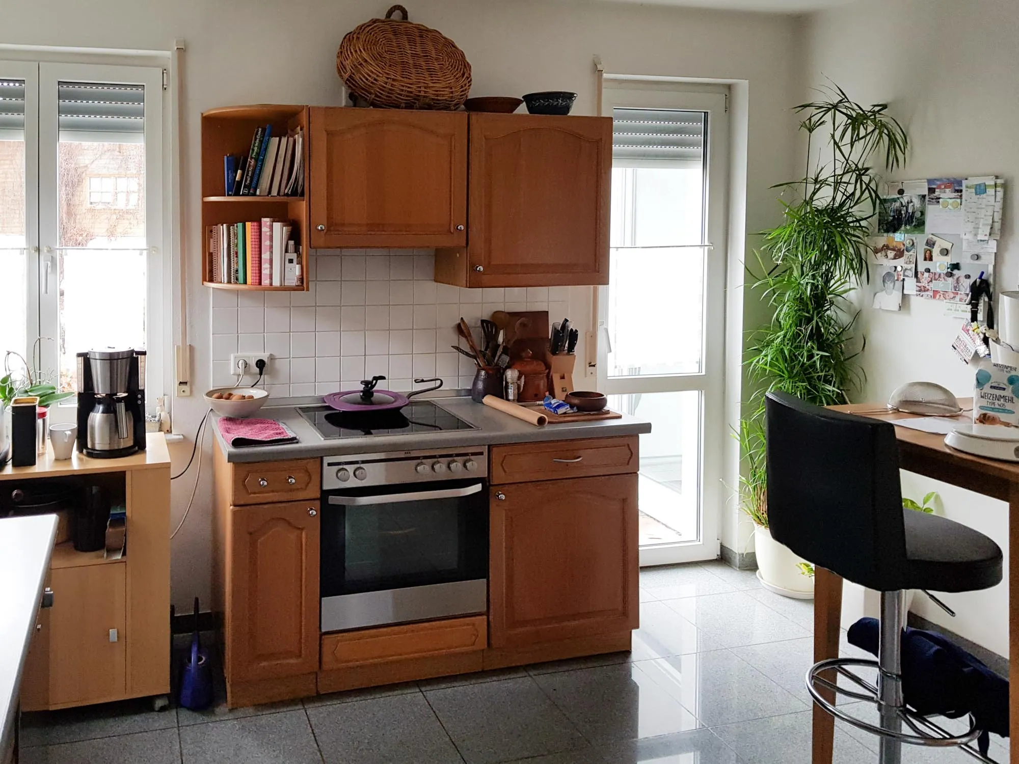 Vorher-Aufnahme einer Bestands-Küche im Landhausstil mit Kassettenfronten aus Eichenholz, Einbau-Backofen mit Edelstahlblende, Kochfeld mit roter Pfanne, weiße Wandfliesen, große Fenster mit Tageslicht, schwarzer Barhocker und Pflanzen im bewohnten Zustand – Ausgangssituation vor der Küchenplanung von Dietmar Kraus bei Broßler Küche Aktiv