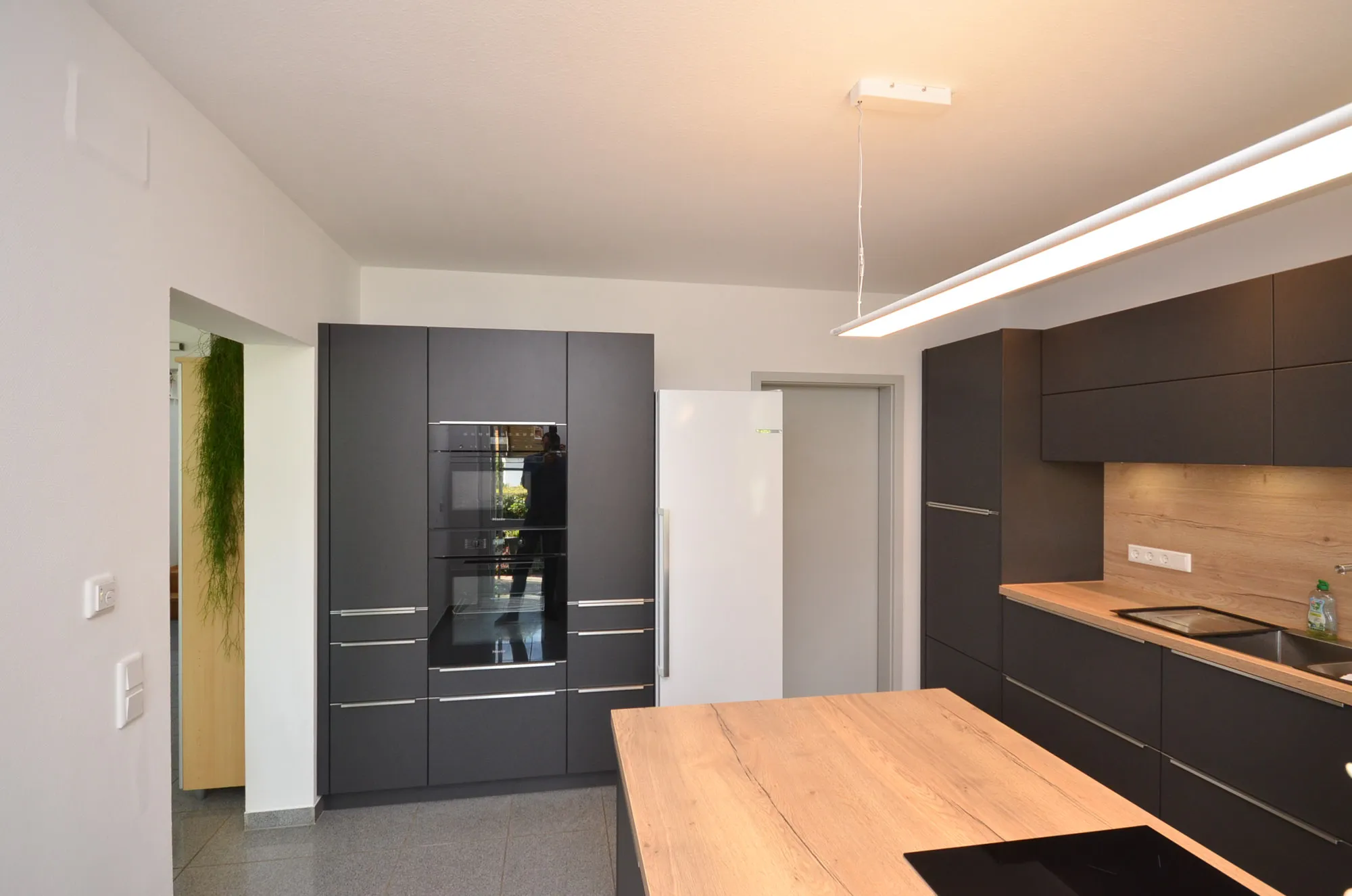 Anthrazitfarbene Küche mit raumhoher Hochschrankzeile, integriertem Backofen und Mikrowelle sowie Side-by-Side-Kühlschrank, gegenüber Arbeitszeile mit Eichen-Holzoptik-Rückwand und Induktionskochfeld, große Kochinsel mit Eichen-Arbeitsplatte vorn, lineare Pendelleuchte und Eichendielen-Boden – Küchenplanung von Dietmar Kraus für Broßler Küche Aktiv