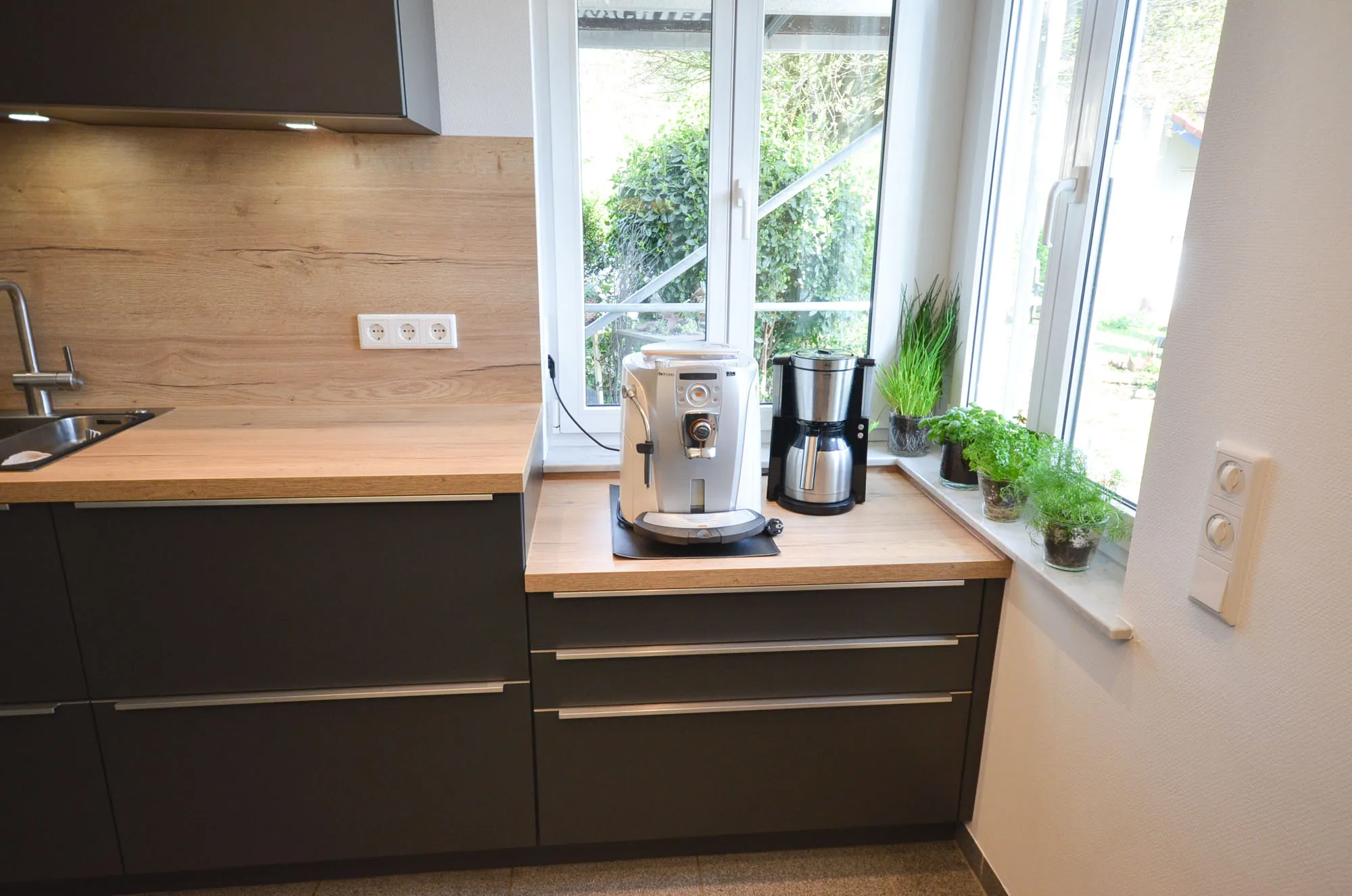 Detailansicht der anthrazitfarbenen Küche am Fenster mit Eichen-Arbeitsplatte und passender Nischenrückwand, Kaffeevollautomat und Filterkanne auf der Arbeitsfläche, Kräutertopf-Reihe auf der Fensterbank, Blick ins Grüne – Küchenplanung von Dietmar Kraus für Broßler Küche Aktiv