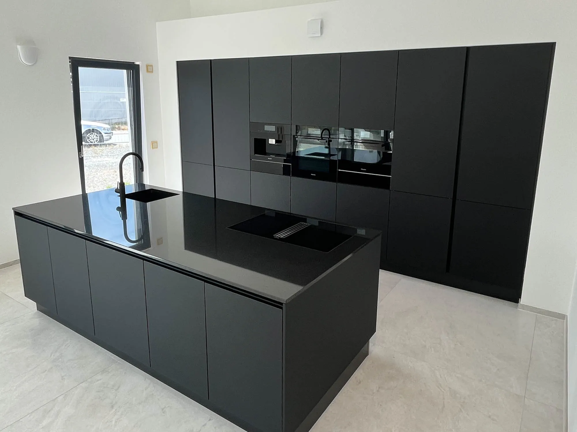 Schwarze grifflose Küche mit großer Kochinsel, schwarzer Steinarbeitsplatte und integriertem Induktionskochfeld mit Muldenlüfter, eingelassene Edelstahl-Spüle mit hoher schwarzer Armatur, im Hintergrund Hochschrankzeile mit Backofen und Mikrowelle, große Fenstertür nach draußen, heller Steinfliesenboden – Küchenplanung von Dietmar Kraus für Broßler Küche Aktiv
