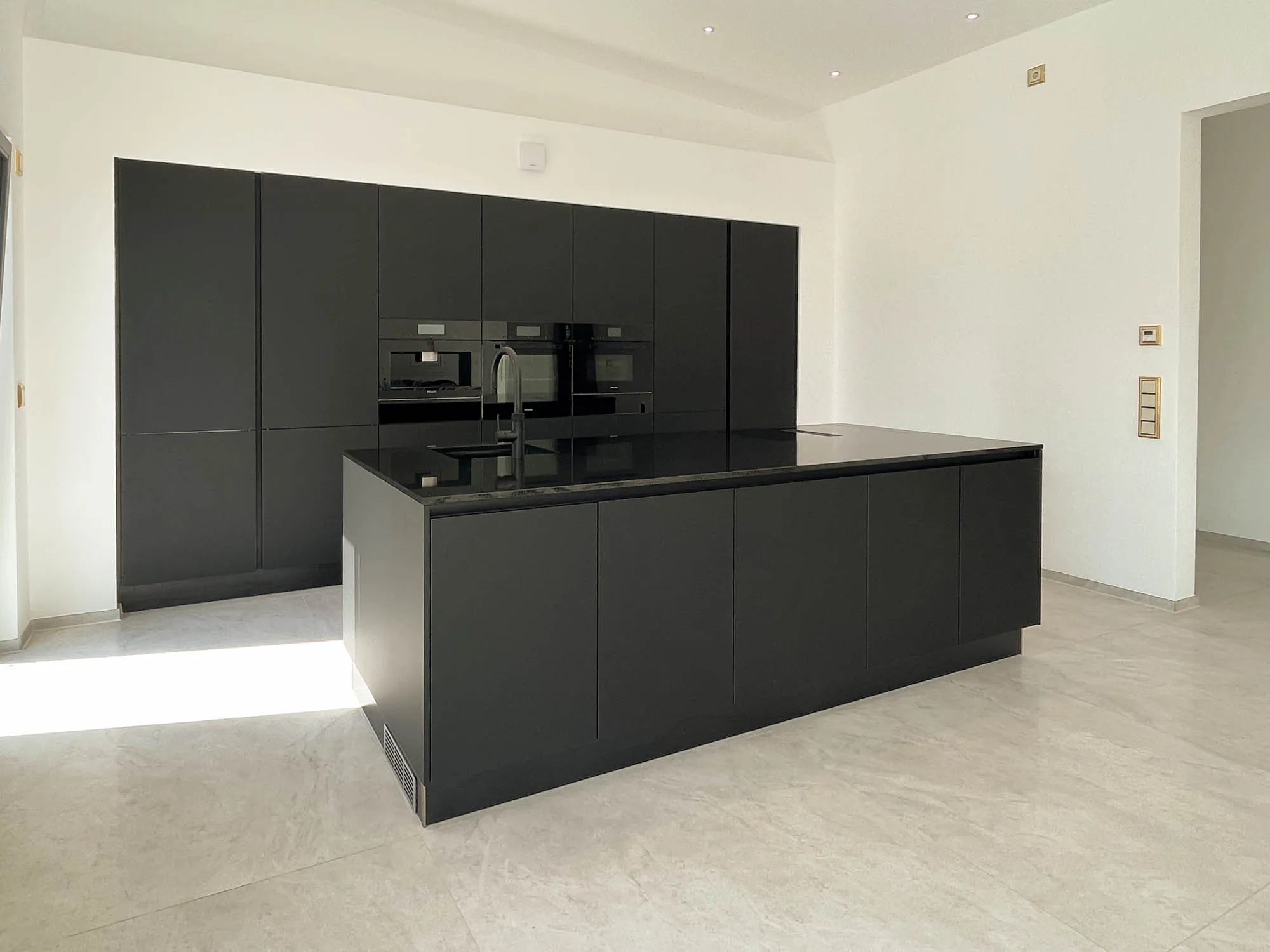Schwarze grifflose Küche mit freistehender Kochinsel und schwarzer Steinarbeitsplatte mit integriertem Induktionskochfeld, im Hintergrund Hochschrankzeile mit Backofen, Mikrowelle und Kaffeevollautomat in Schwarz, heller Steinfliesenboden – Küchenplanung von Dietmar Kraus für Broßler Küche Aktiv