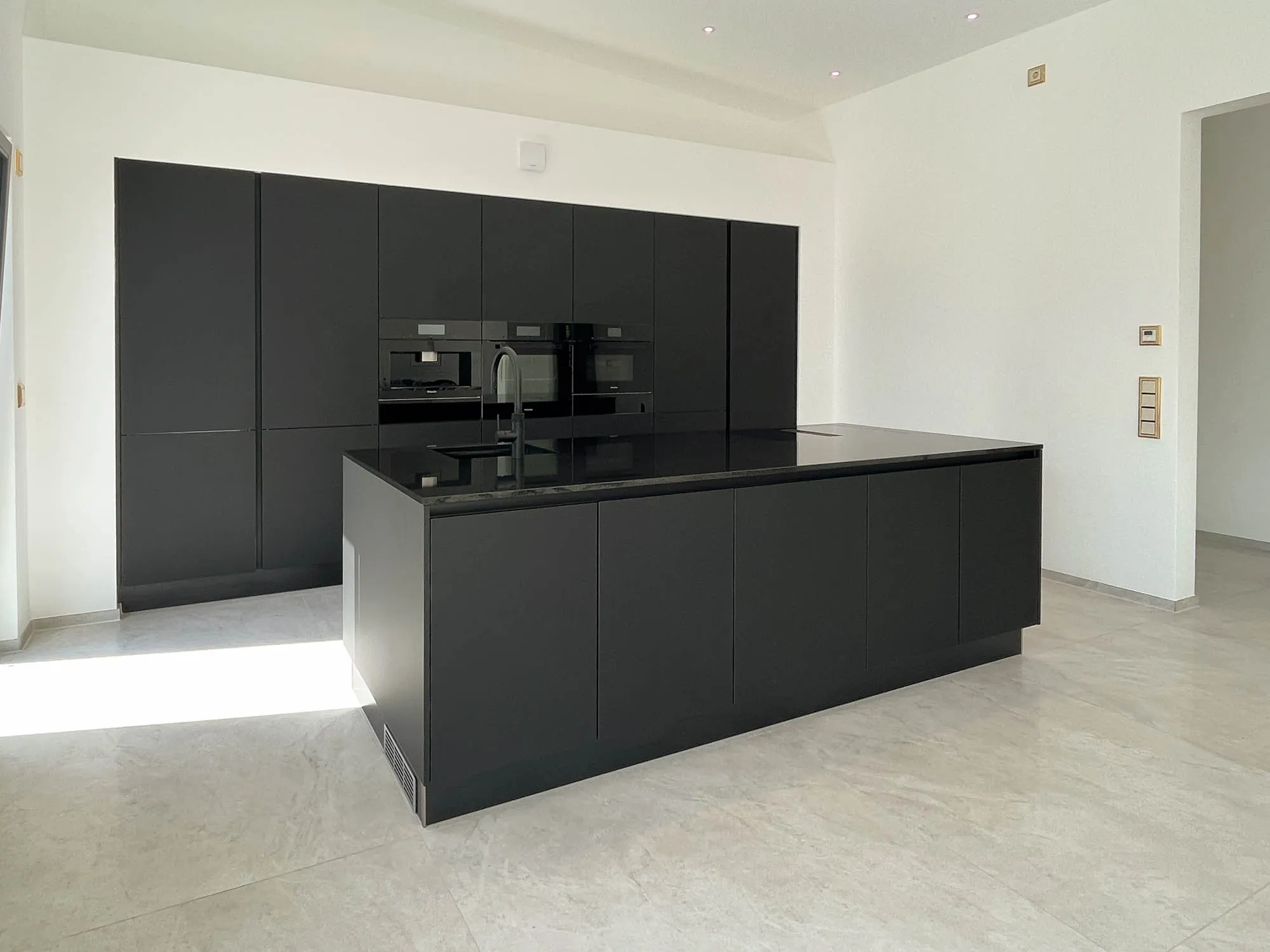 Schwarze grifflose Küche mit freistehender Kochinsel und schwarzer Steinarbeitsplatte mit integriertem Induktionskochfeld, im Hintergrund Hochschrankzeile mit Backofen, Mikrowelle und Kaffeevollautomat in Schwarz, heller Steinfliesenboden – Küchenplanung von Dietmar Kraus für Broßler Küche Aktiv