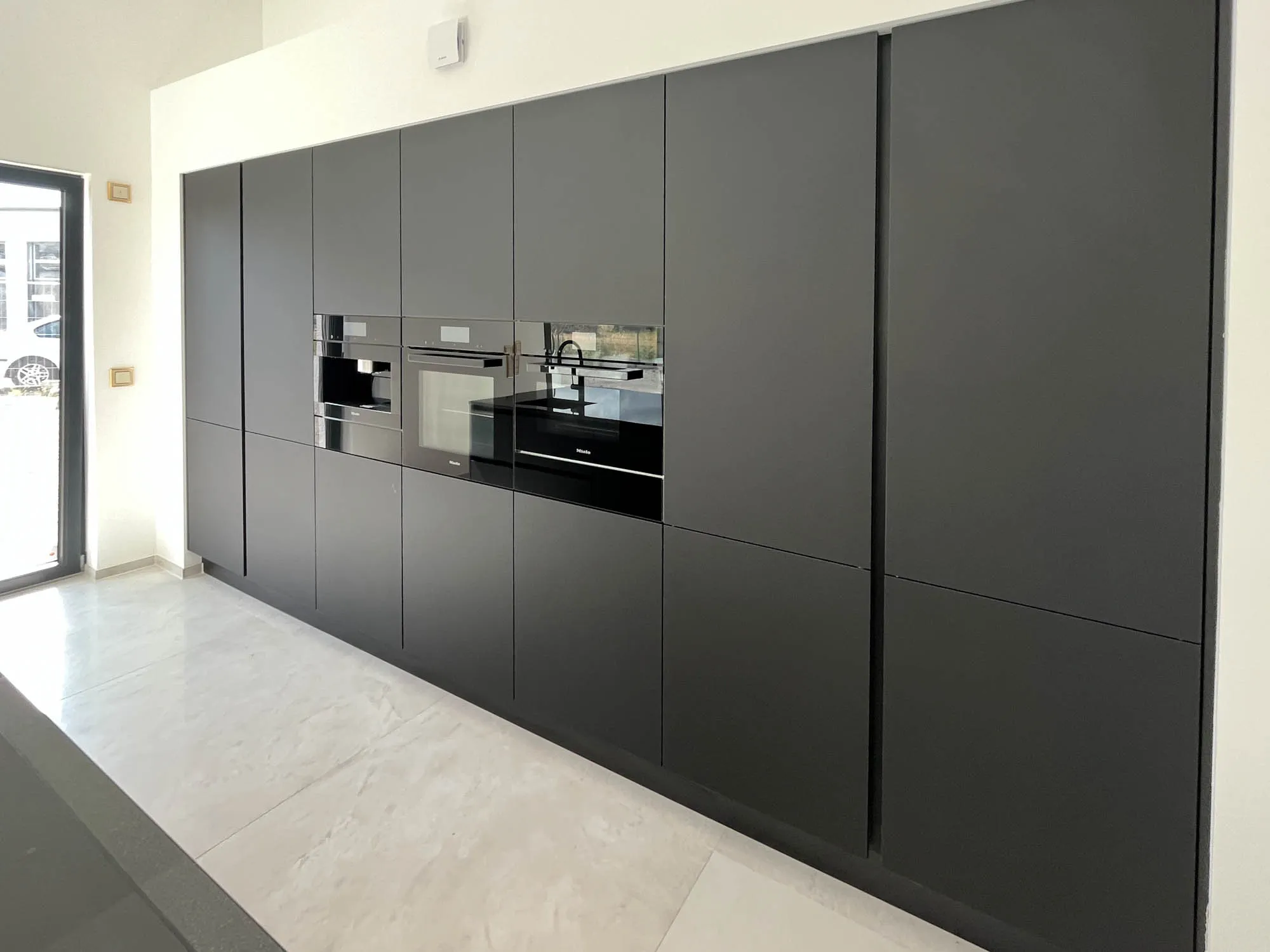 Lange schwarze Hochschrankzeile in mattem Anthrazit-Schwarz mit grifflosen Fronten, integriertem Backofen und Mikrowelle mittig, große Fenstertür links mit Tageslicht, heller Steinfliesenboden – Küchenplanung von Dietmar Kraus für Broßler Küche Aktiv