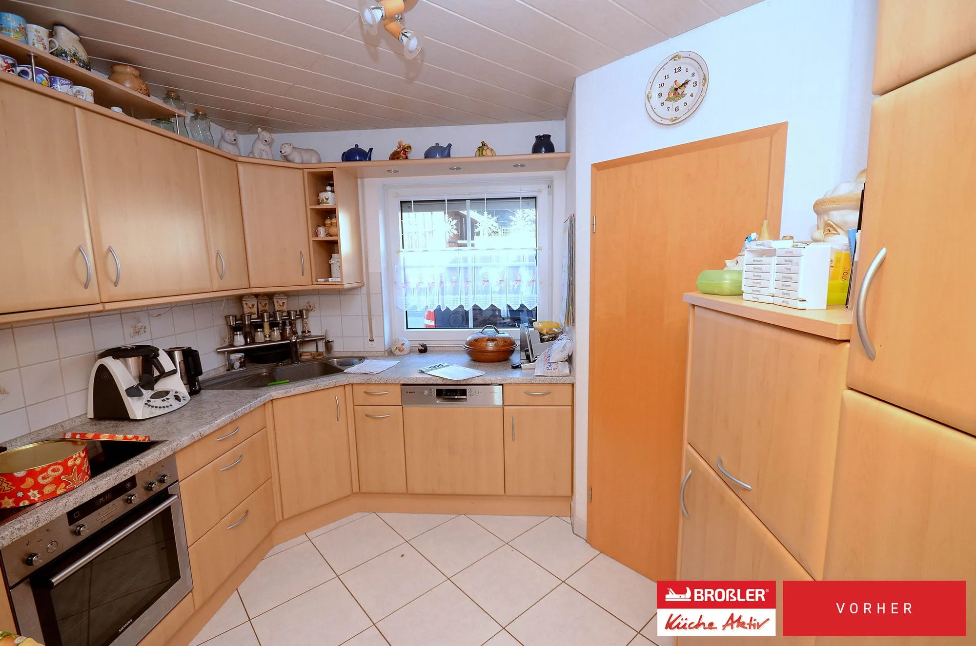 Vorher-Aufnahme einer U-Küche in heller Buche-Holzoptik mit Spüle unter Sprossenfenster, Backofen und weißen Wandfliesen.