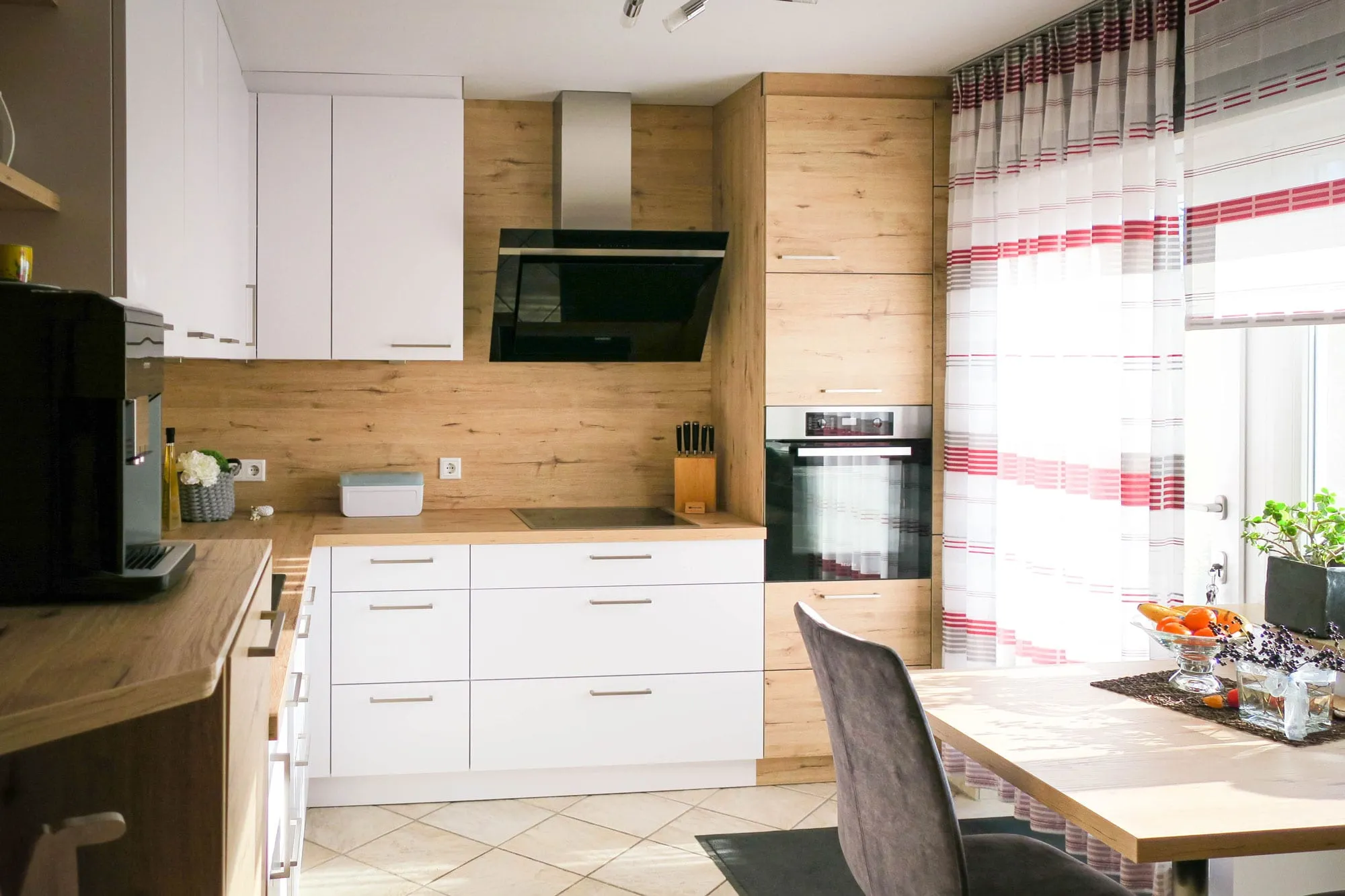 Gesamtansicht der Wohnküche: Kochbereich mit Wand-Dunstabzug, Backofen-Hochschrank, weißen Fronten und Holz-Rückwand, im Vordergrund Esstisch mit grauem Polsterstuhl
