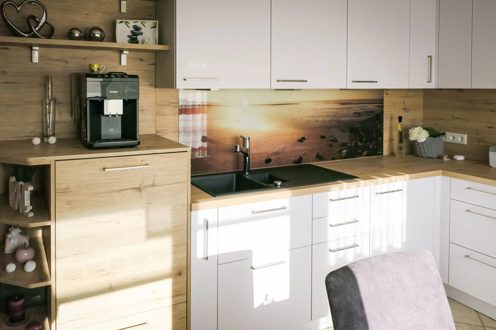 Helle L-Küche mit weißen grifflosen Fronten, Holz-Arbeitsplatte, Spüle mittig und fotorealistischem Spritzschutz mit Sonnenuntergang-Motiv, links Eckschrank mit offenem Holzregal