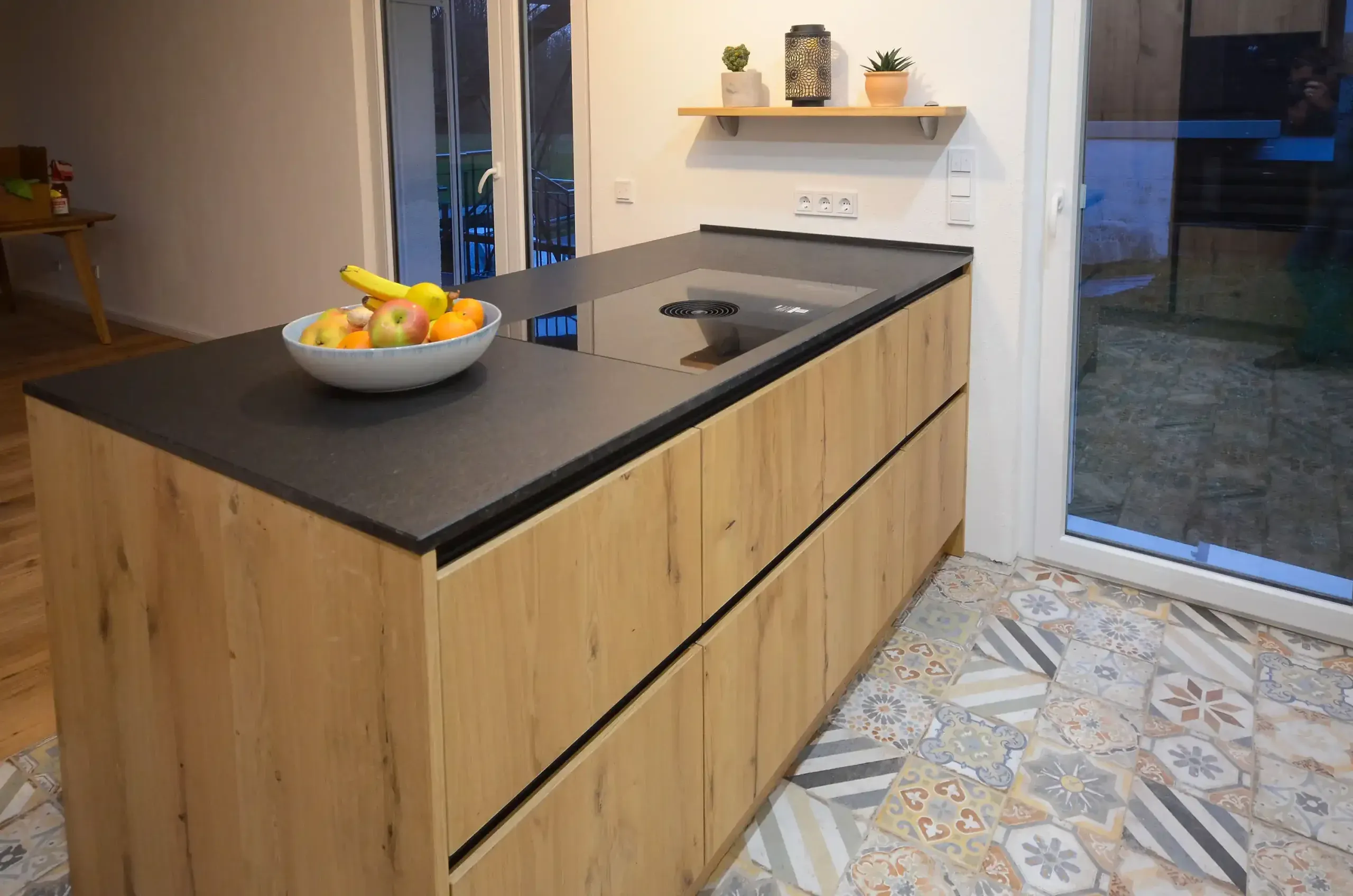 Detailansicht einer Kochinsel mit Wildeiche-Fronten, dunkler Arbeitsplatte, integriertem Kochfeld, Obstschale und Wandregal mit Pflanzen, Glasschiebetür rechts und gemustertem Patchwork-Fliesenboden – Referenzprojekt von Broßler Küche Aktiv (Standort Ehrbach/Großheubach)