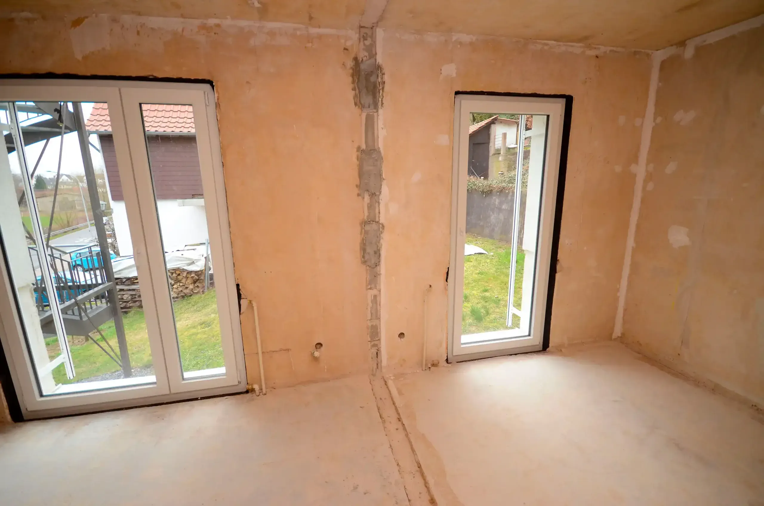 Eckraum im Rohbau mit zwei bodentiefen Fenstertüren zur Terrasse, gespachtelten Wänden, freiliegender Elektroleitung und Estrichboden – vor der Modernisierung durch Broßler Küche Aktiv (Standort Ehrbach/Großheubach)