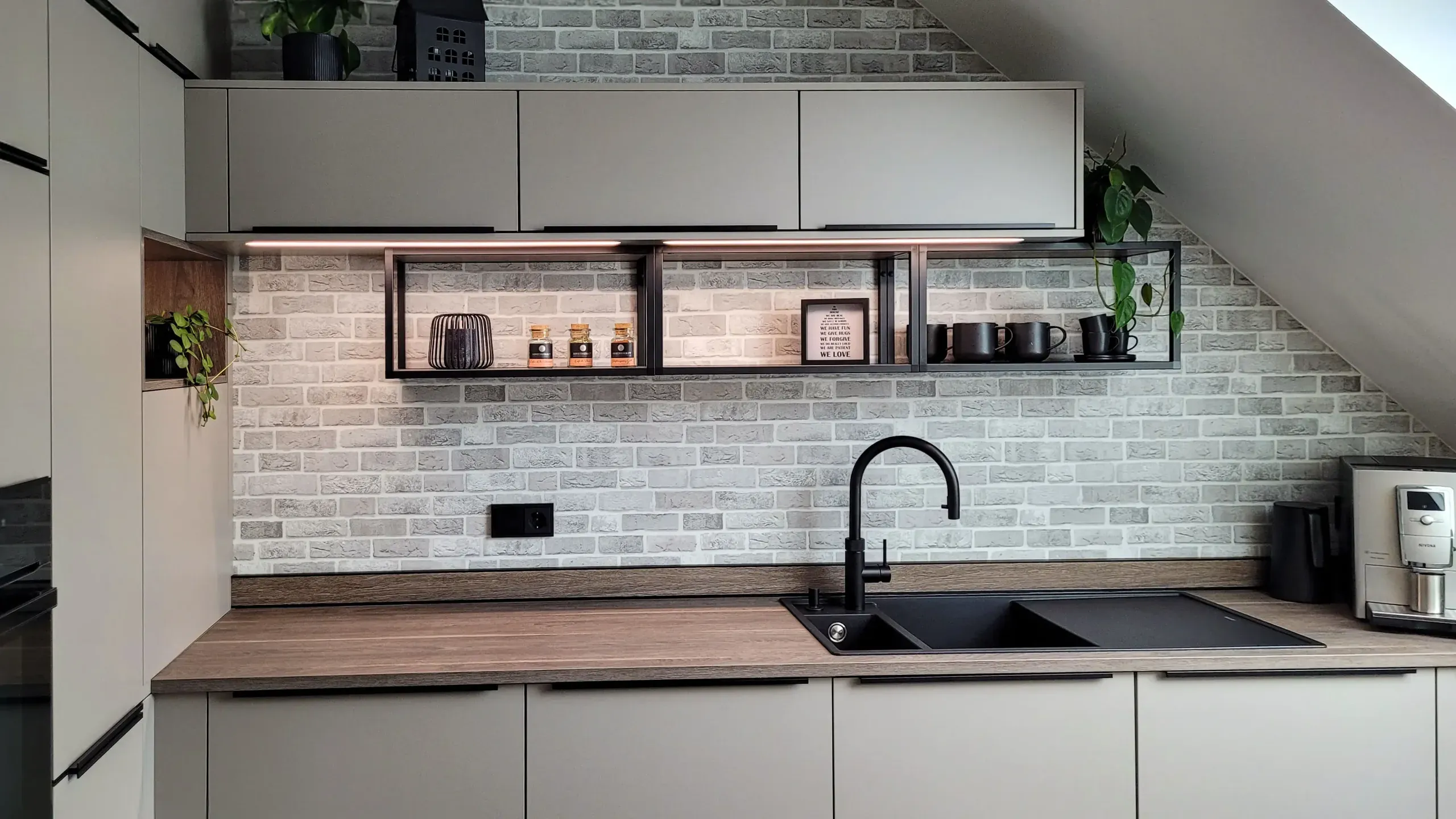 Frontale Detailansicht der Spülzone mit schwarzer Granitspüle, schwebendem Holzregal und Backsteinwand.