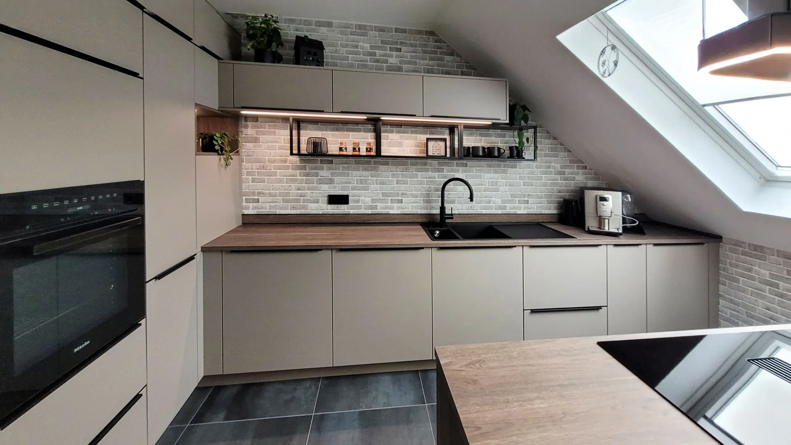 Wandzeile mit Backofen-Hochschrank, Spüle unter Dachfenster und schwebenden Holzregalen vor Backsteinwand.