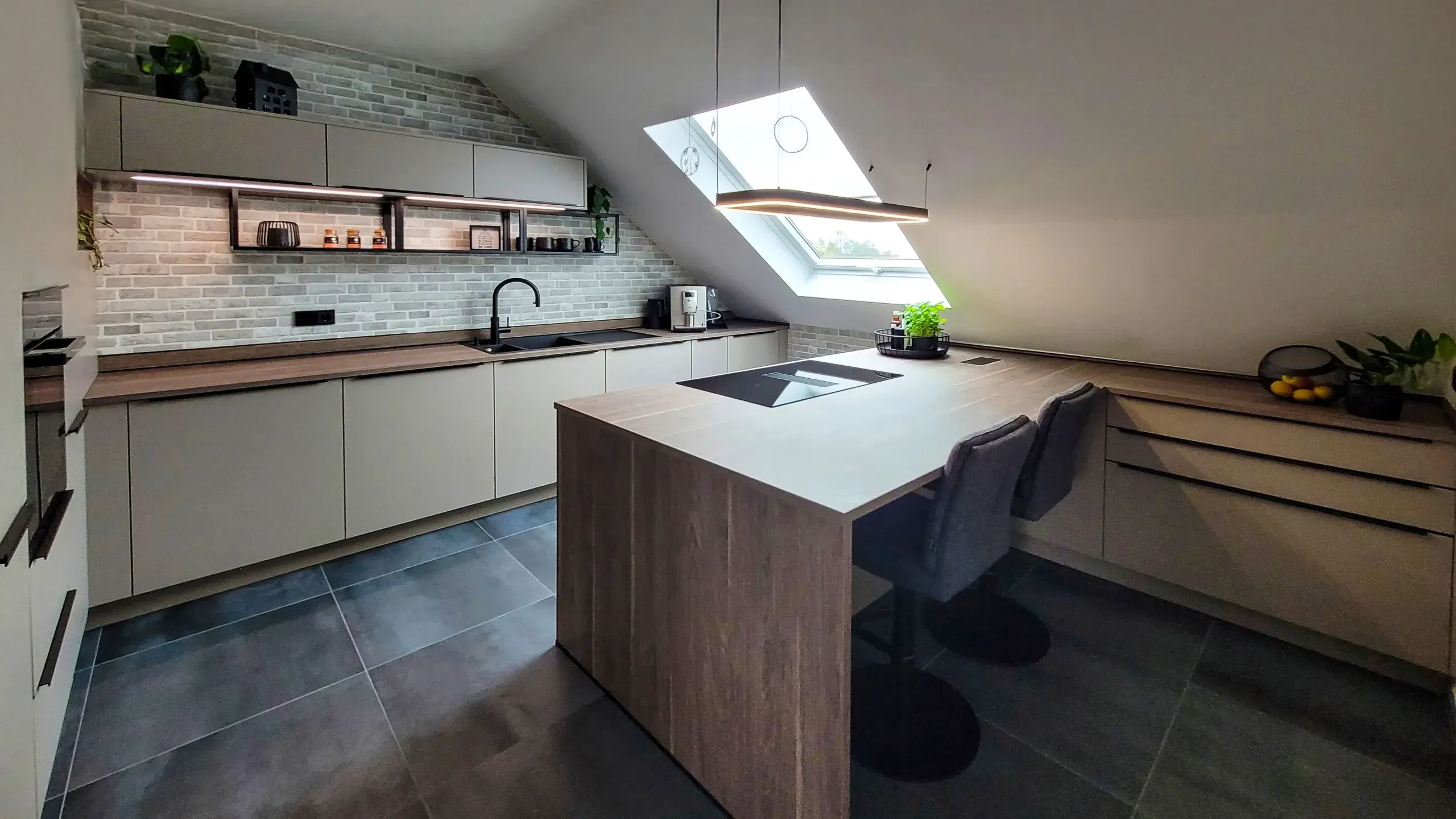 Offene Küche mit Holz-Kochinsel, Dachfenstern und anthrazit-matter Wandzeile vor Backsteinwand.